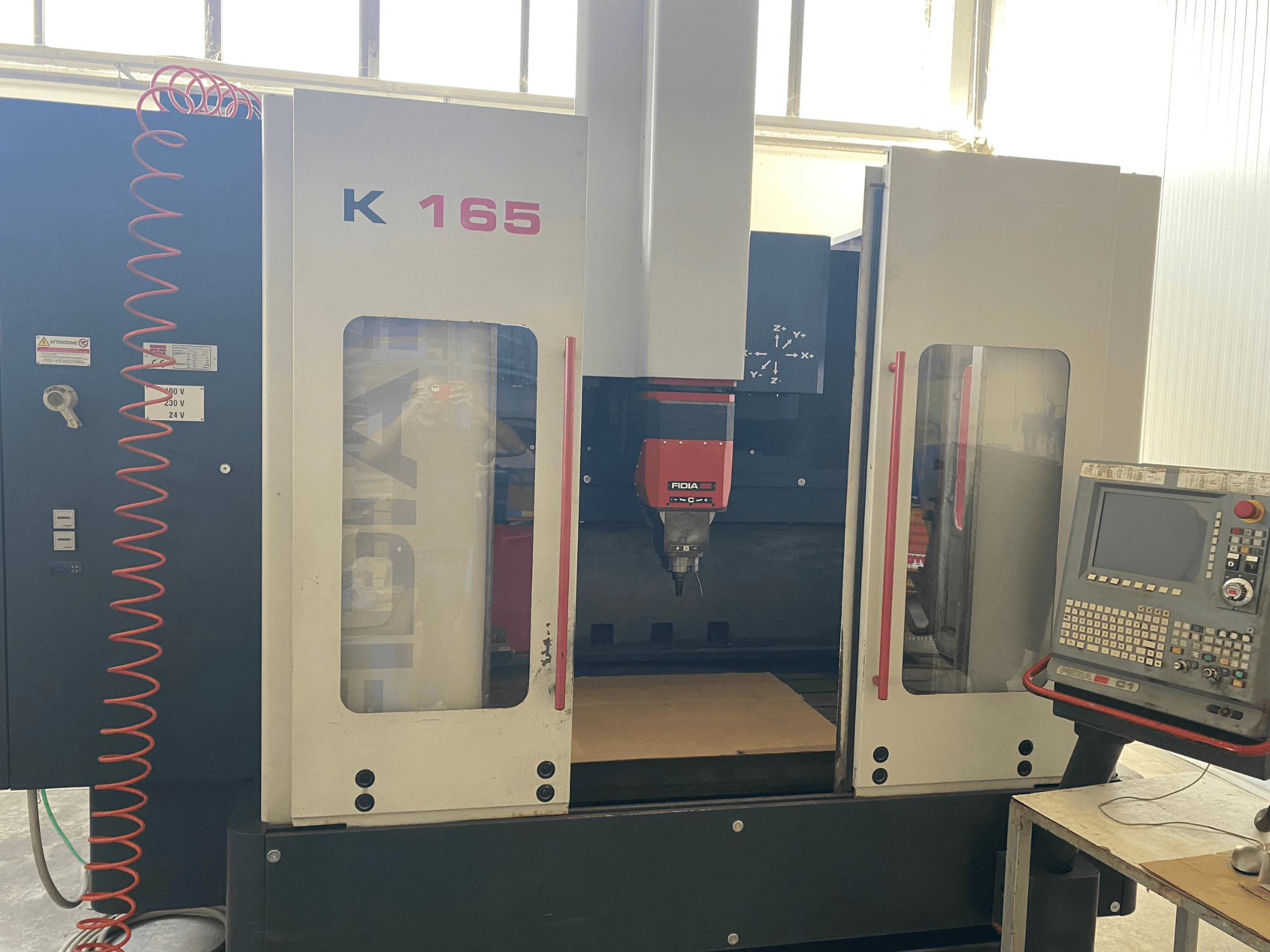 Máquina CNC Fidia K 165 com fuso vermelho e painel de controlo, vista de frente, numa oficina bem iluminada.