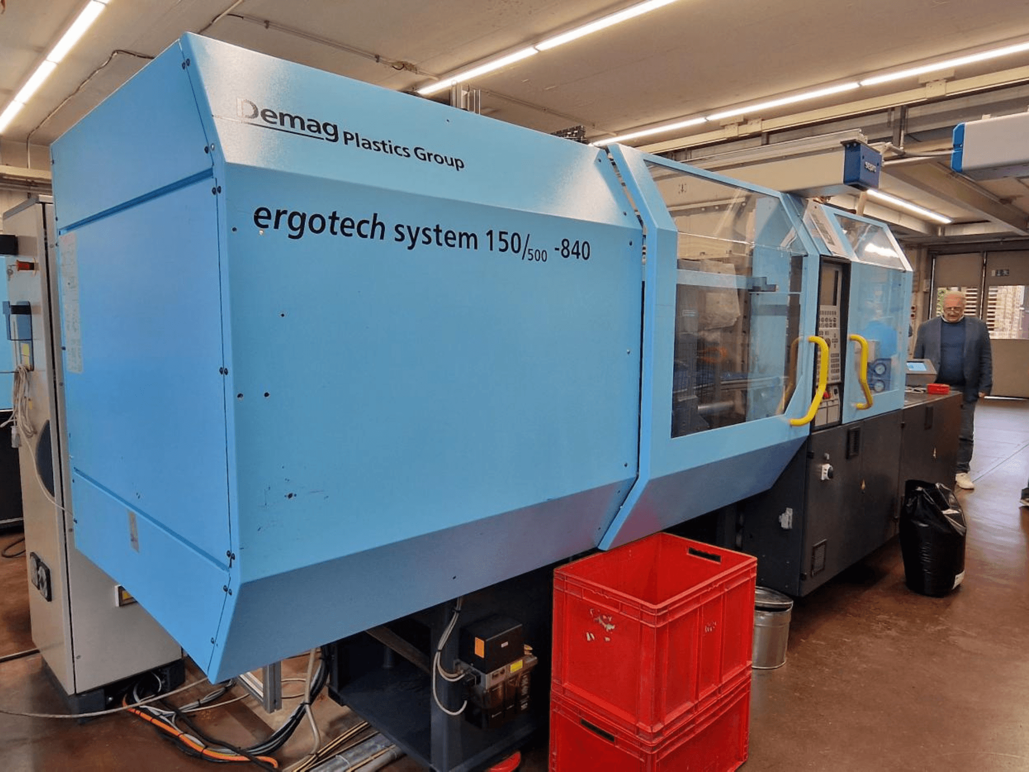 Máquina de moldagem por injeção Demag Ergotech System 150/500-840 em azul, vista lateral, com caixas e operador em segundo plano.