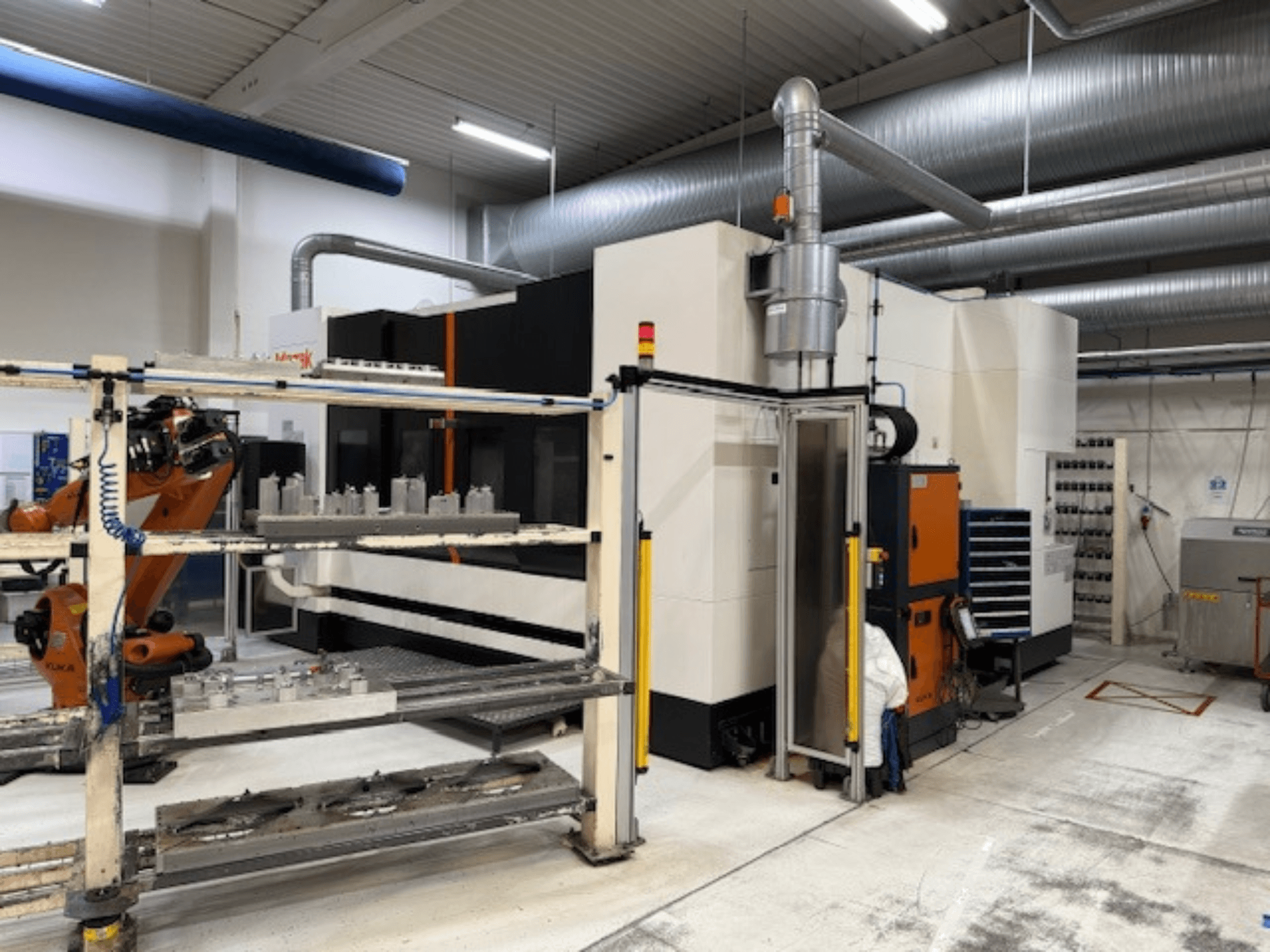 Máquina CNC industrial com braço robótico e barreiras de segurança, com uma vista frontal da configuração num espaço de trabalho arejado.