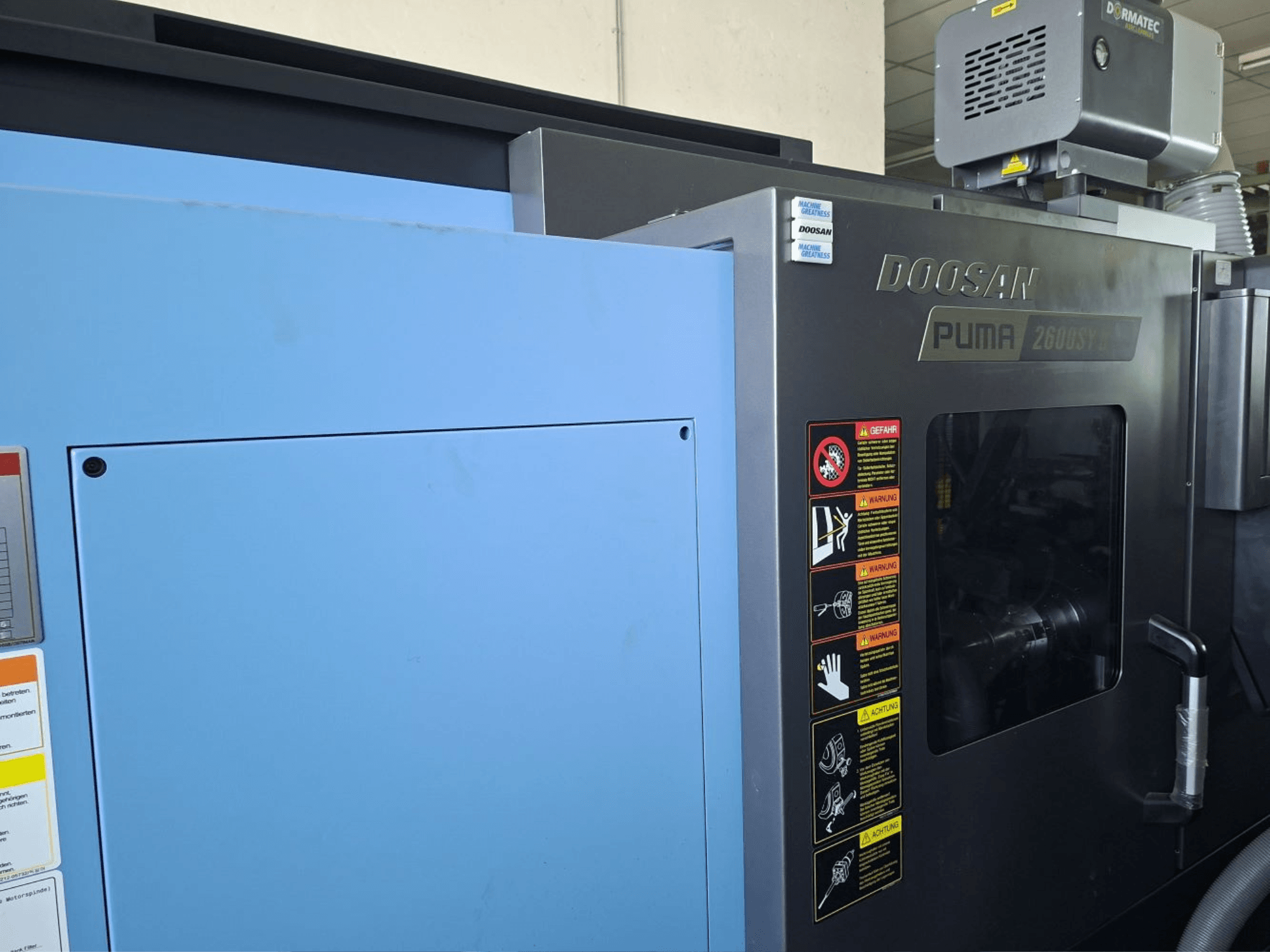 Torneiro horizontal DOOSAN PUMA 2600 SY II, vista frontal, mostrando as etiquetas de segurança e os painéis azuis.