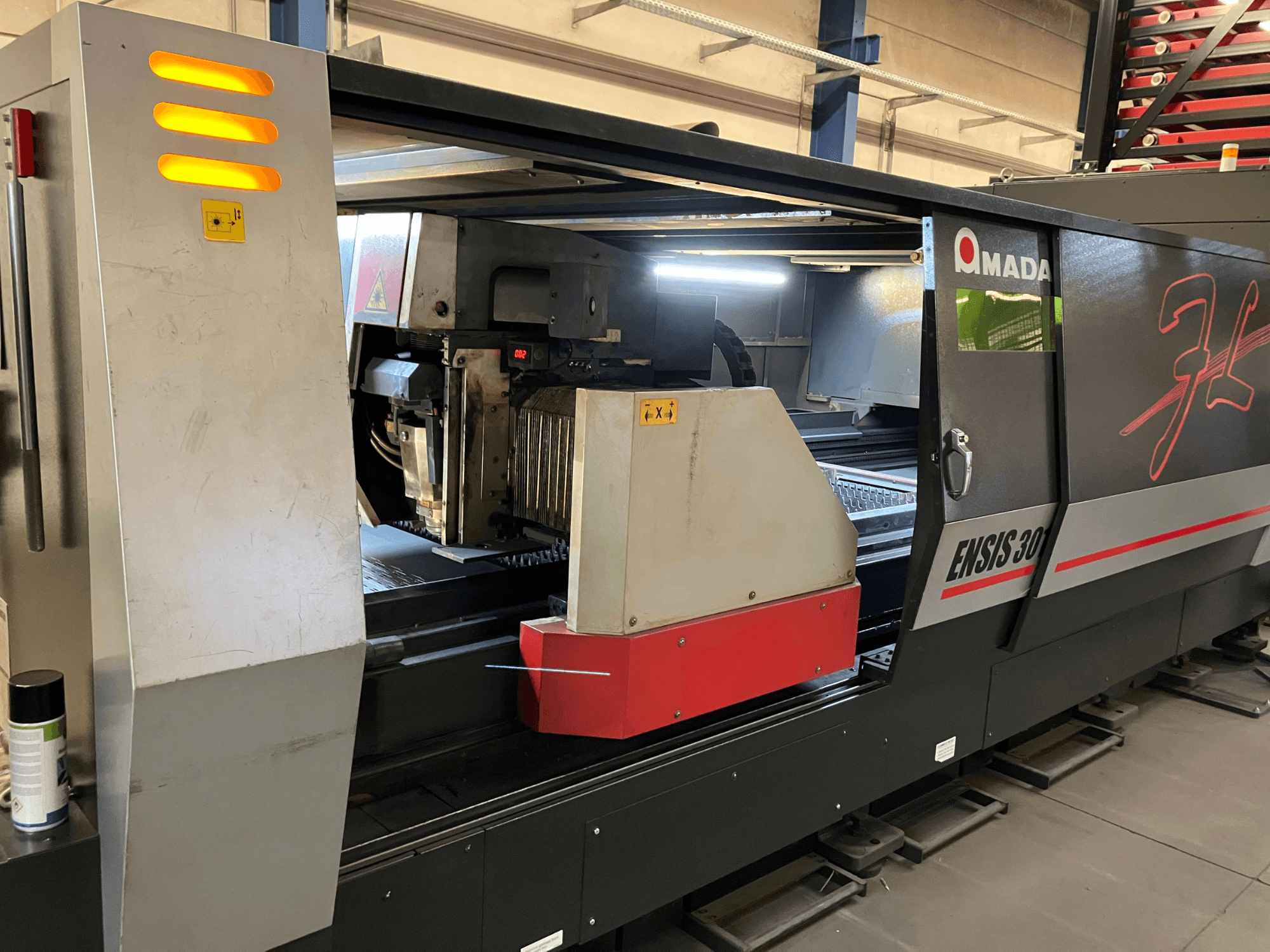 Máquina de corte a laser de fibra AMADA ENSIS 3015 AJ, vista frontal, mostrando a cabeça de corte e os controlos de funcionamento dentro de uma estrutura metálica.
