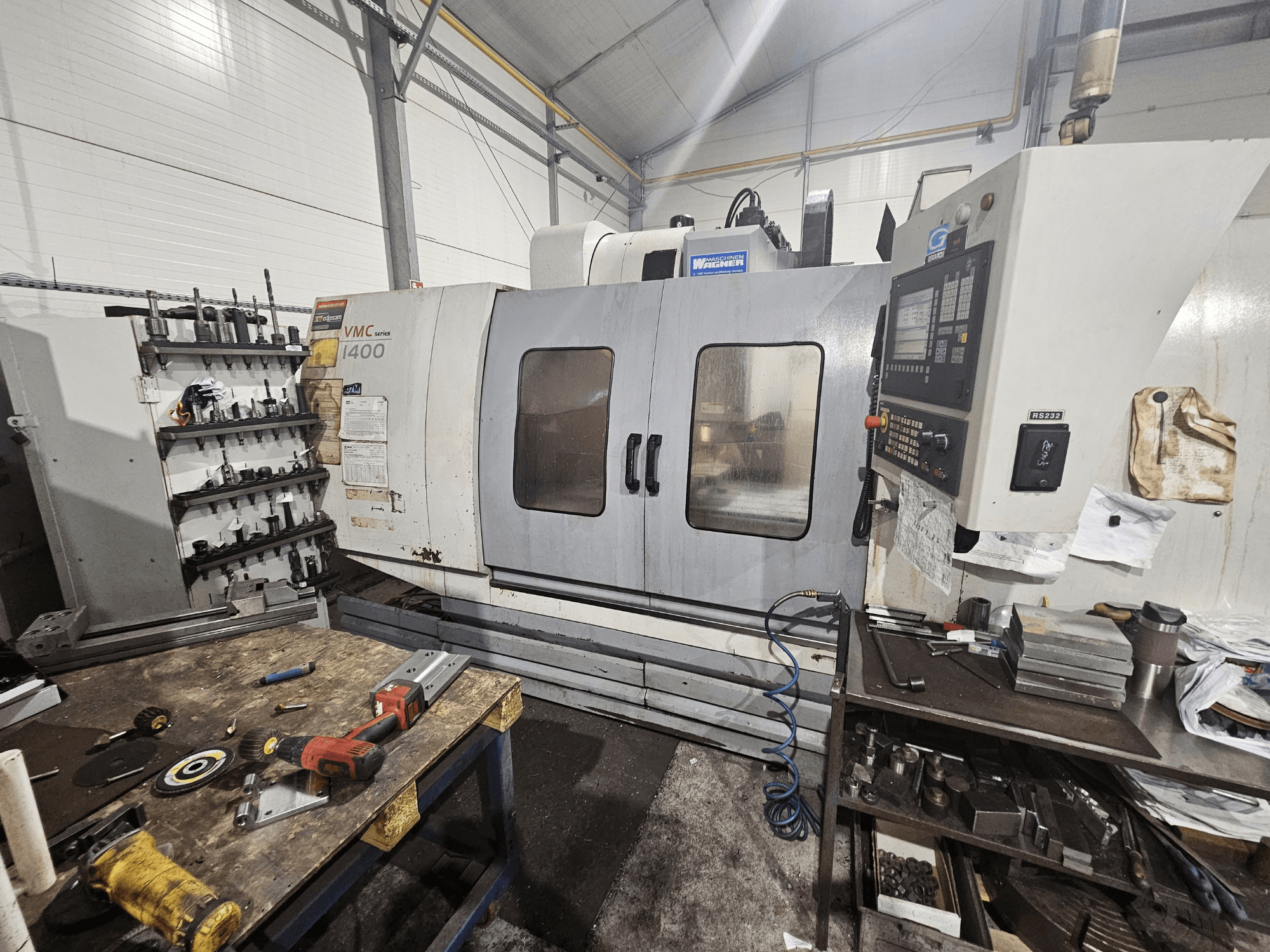 Fresadora VMC 1400 CNC numa oficina, vista frontal; ferramentas e materiais espalhados na bancada de trabalho nas proximidades.