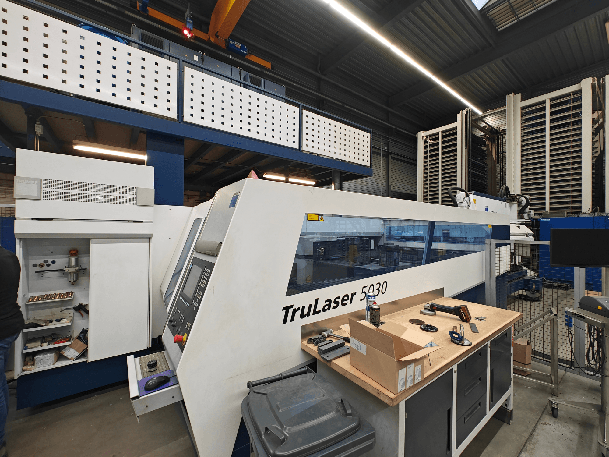 Máquina TruLaser 5030 numa oficina, vista frontal com painel de controlo visível, rodeada de ferramentas e equipamento.