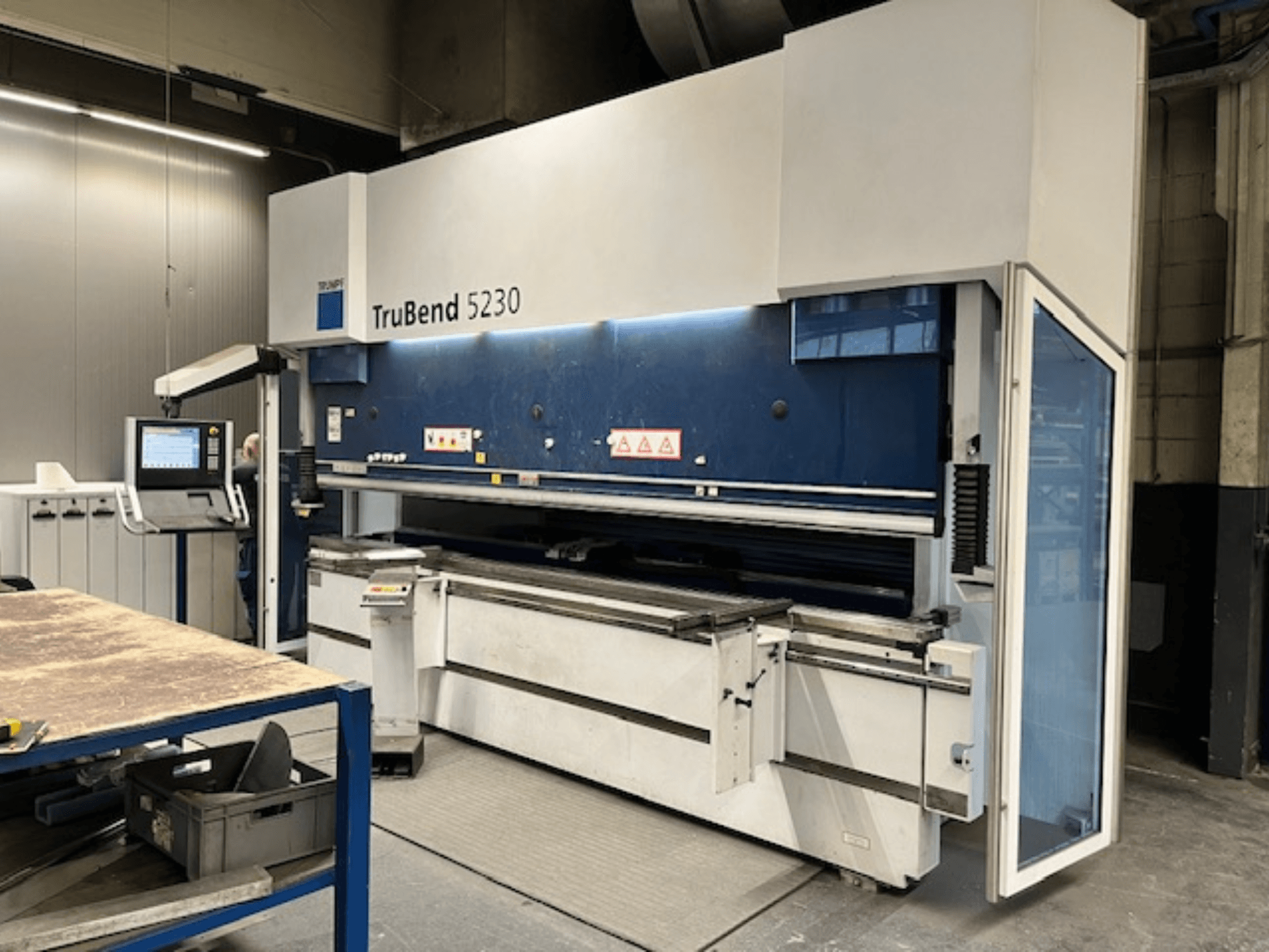 Máquina de dobragem TruBend 5230 em ambiente industrial; vista frontal mostrando o painel de controlo, as caraterísticas de segurança e a mesa de trabalho.