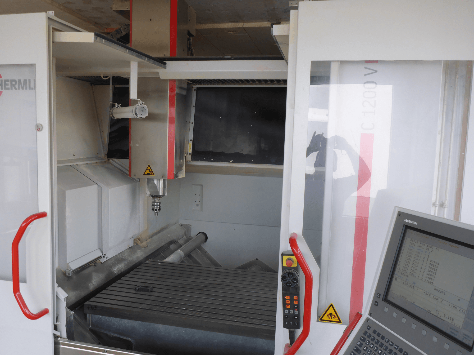 Interior do centro de maquinação vertical HERMLE C 1200 mostrando o porta-ferramentas, o painel de controlo e a mesa de trabalho a partir de uma vista frontal.