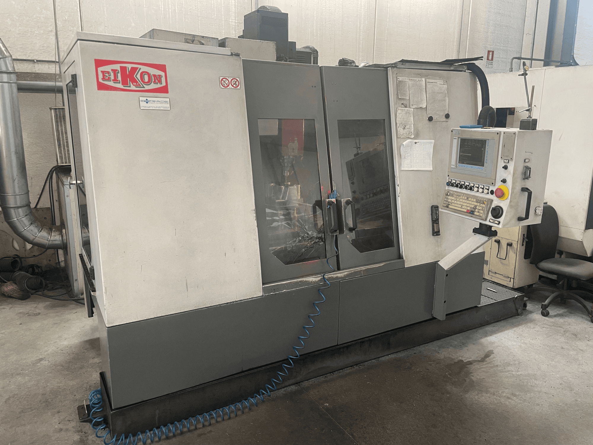 Máquina CNC EIKON vista de frente, com um painel de controlo, grandes portas de vidro e uma mangueira de ar azul no chão.