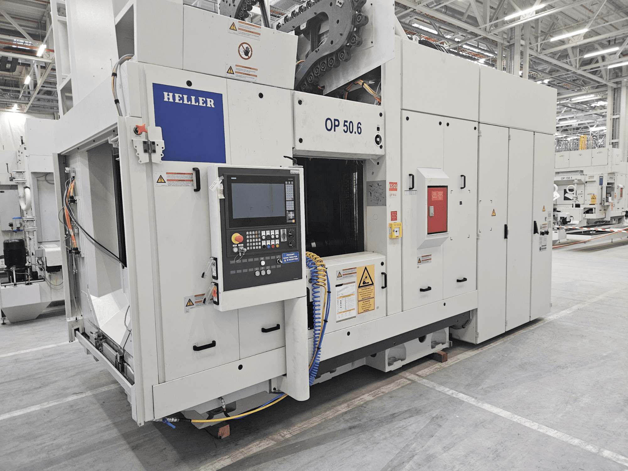 Centro de maquinação Heller OP 50.6, vista frontal, com um painel de controlo, etiquetas de aviso e mangueiras visíveis num ambiente de fábrica.