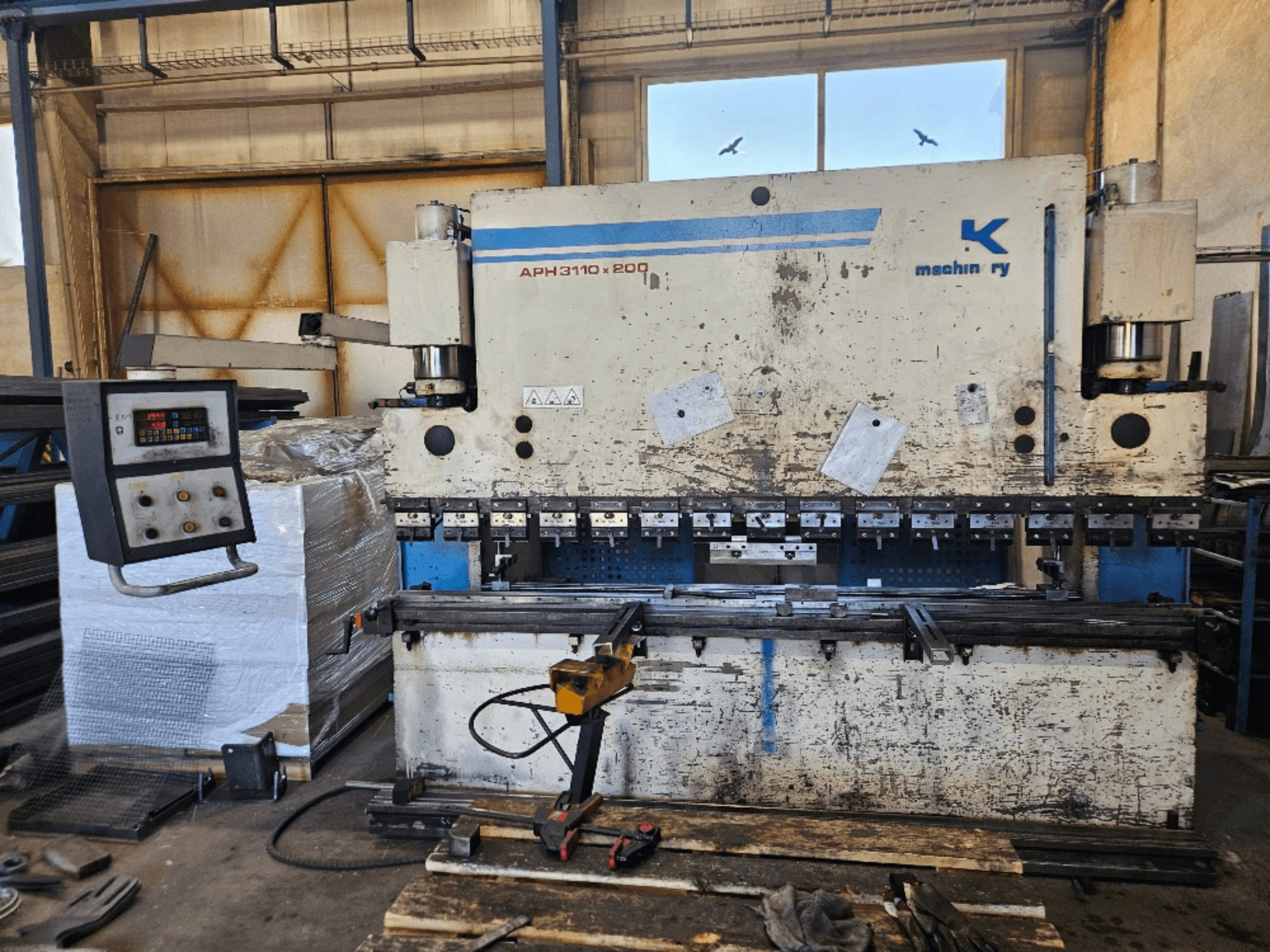 Prensa dobradeira Baykal APH 3110x200 numa oficina, vista frontal, com painel de controlo, chapas metálicas e ferramentas visíveis.