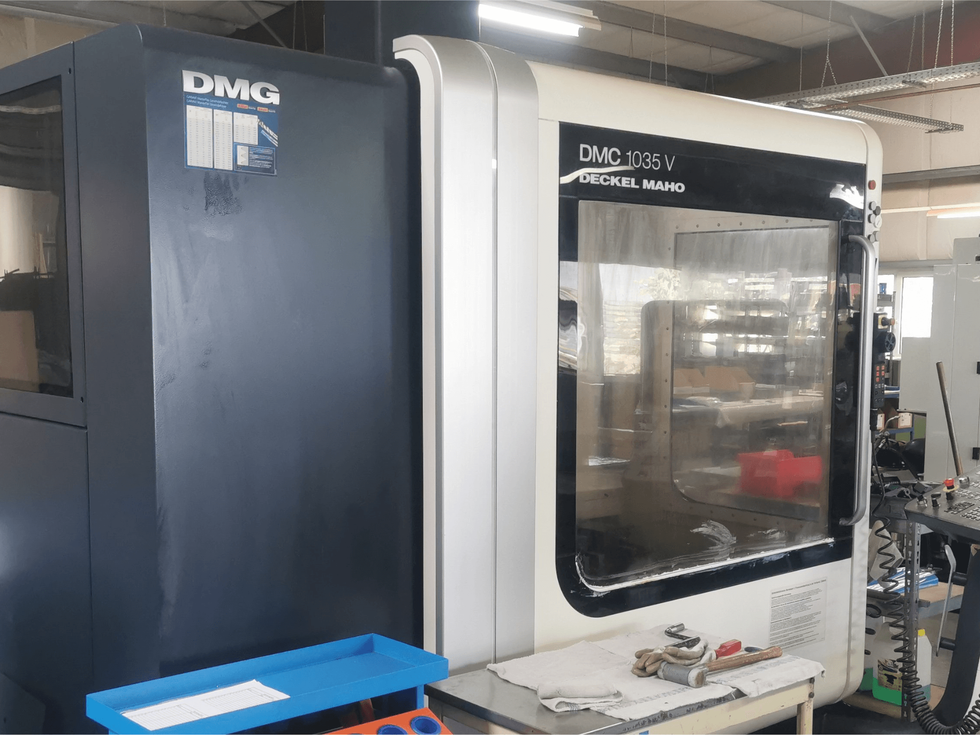 Fresadora DMG DMC 1035 V numa oficina, vista frontal com tabuleiro de ferramentas azul e luvas na superfície.