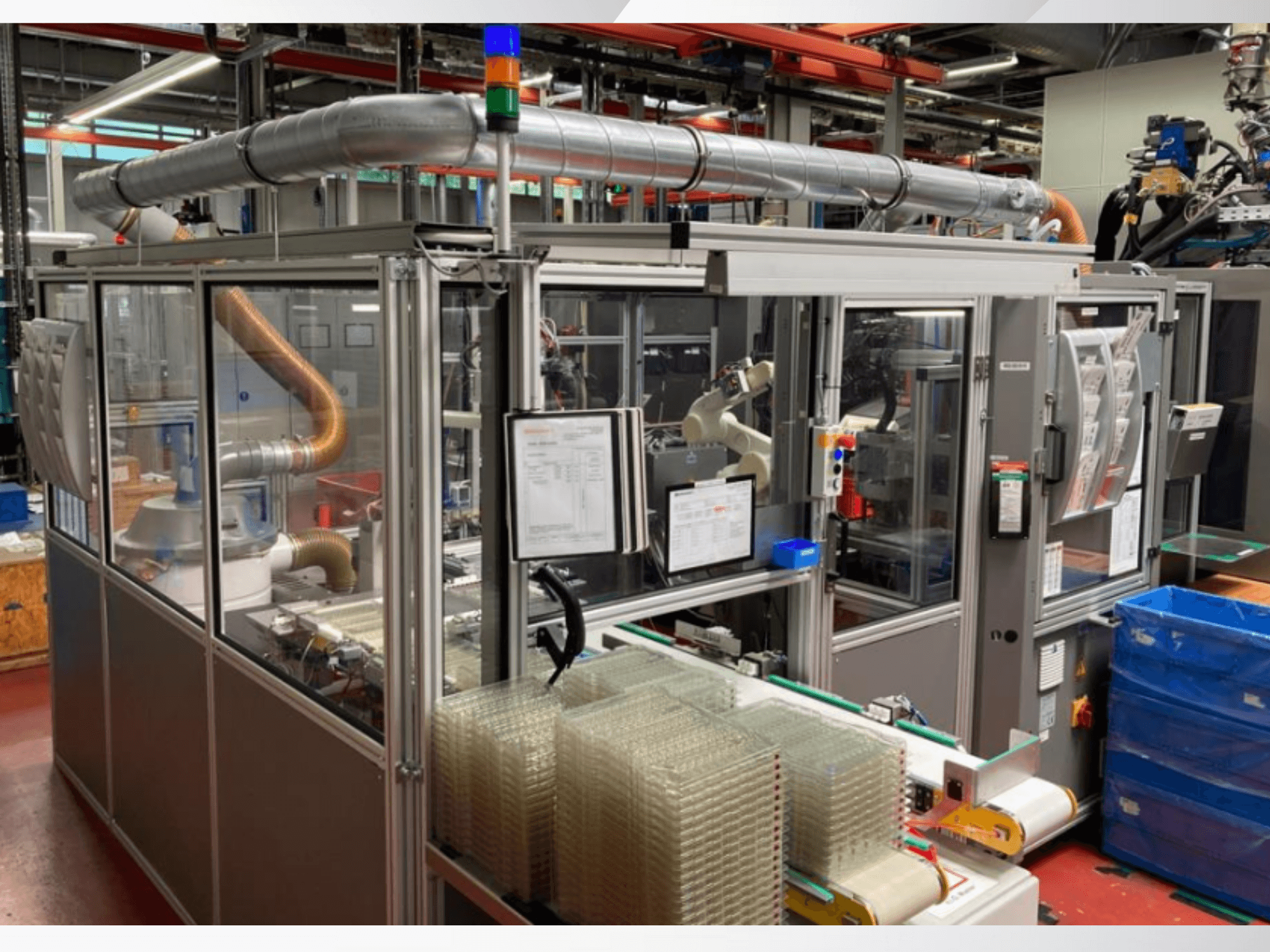 Máquina de montagem automatizada com braço robótico, painéis laterais transparentes e contentores empilhados de artigos transparentes, vista frontal.