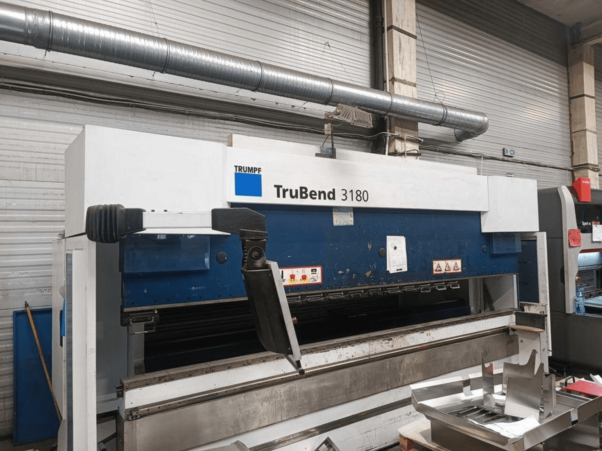 Máquina de dobragem Trumpf TruBend 3180 numa oficina, vista lateral mostrando o painel de controlo e as peças de metal na mesa.