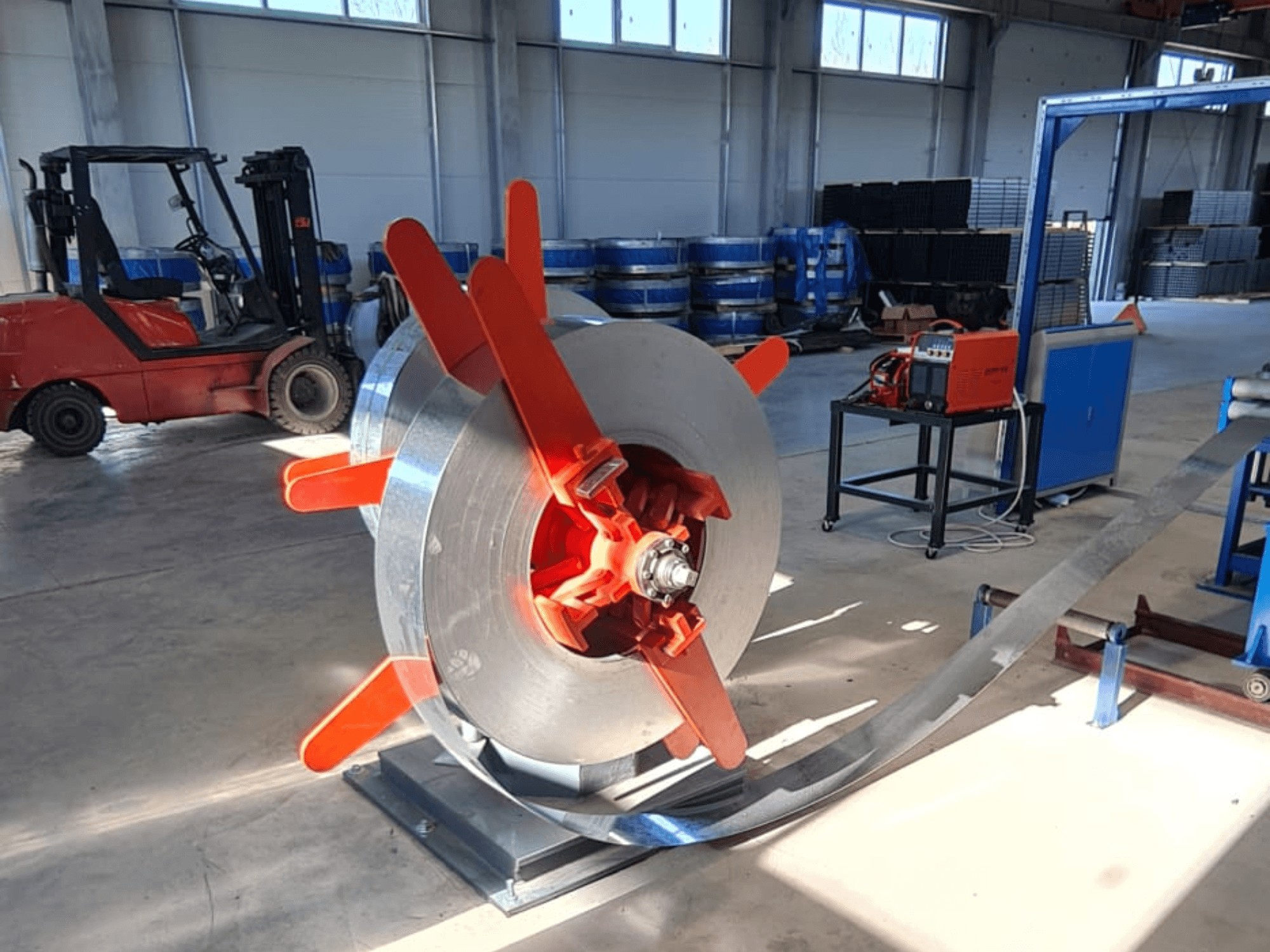 Bobina de metal com pegas vermelhas num suporte, vista de um ângulo. Fundo industrial com maquinaria e um empilhador.