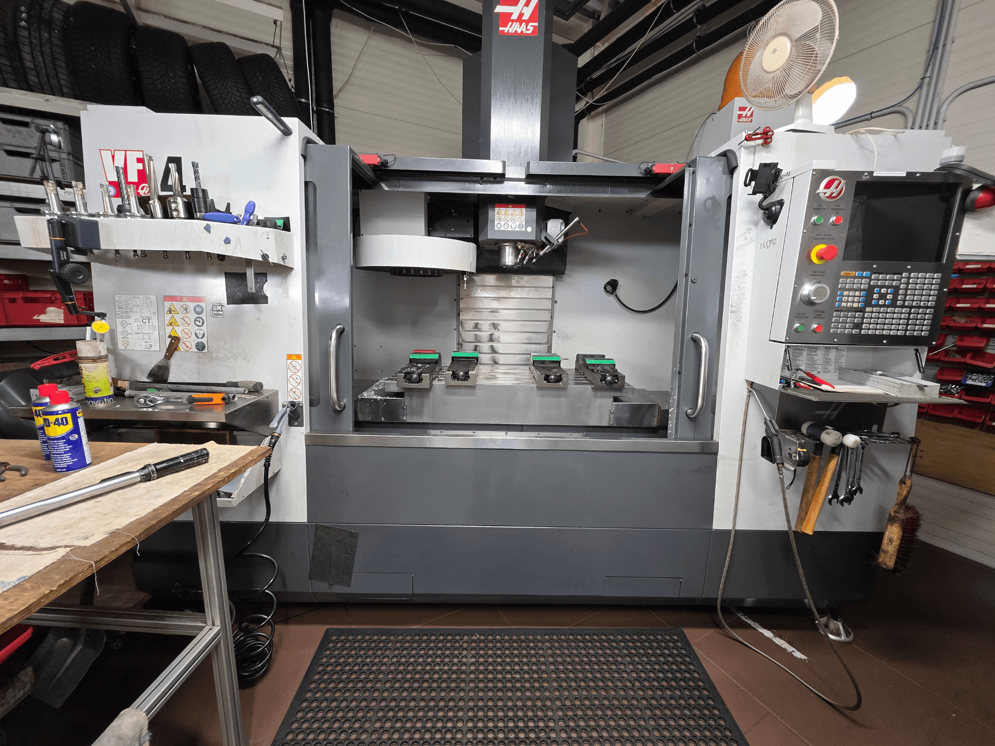 Vista frontal de uma fresadora CNC Haas VF-4 numa oficina, com ferramentas, uma interface de computador e duas peças de trabalho na mesa.