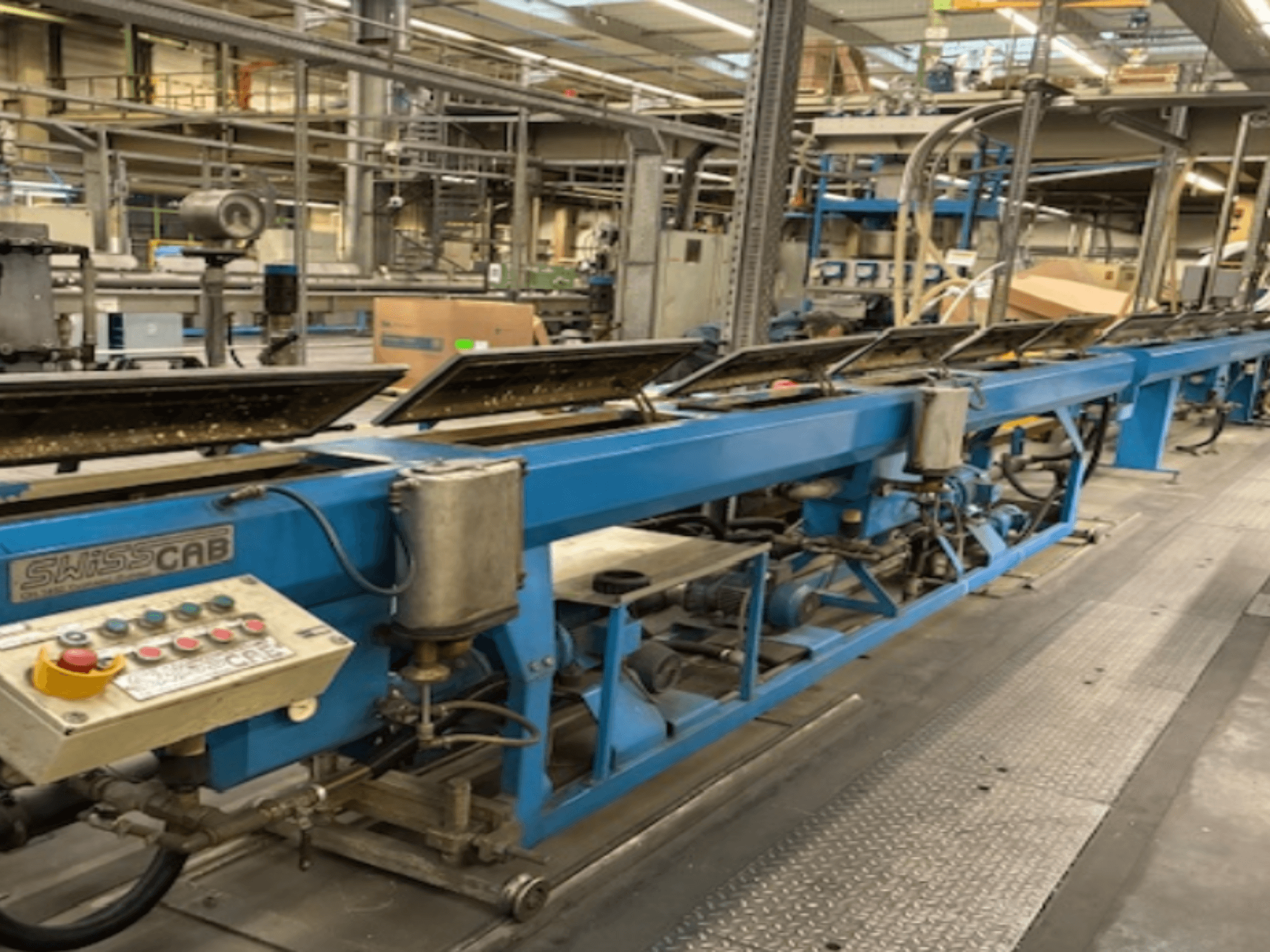 Máquina industrial SwissCab azul num ambiente de fábrica, com botões de funcionamento e um sistema de transporte, vista lateral.