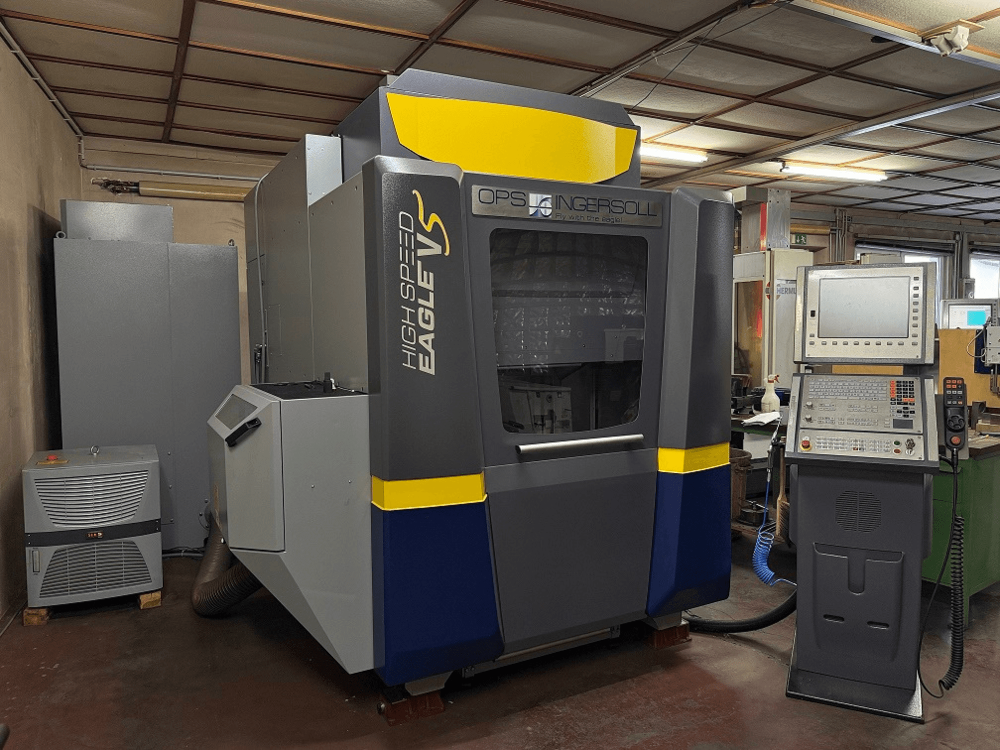 Máquina Eagle V de alta velocidade da OPS Ingersoll, vista lateral, com painel de controlo e equipamento adjacente em ambiente industrial.