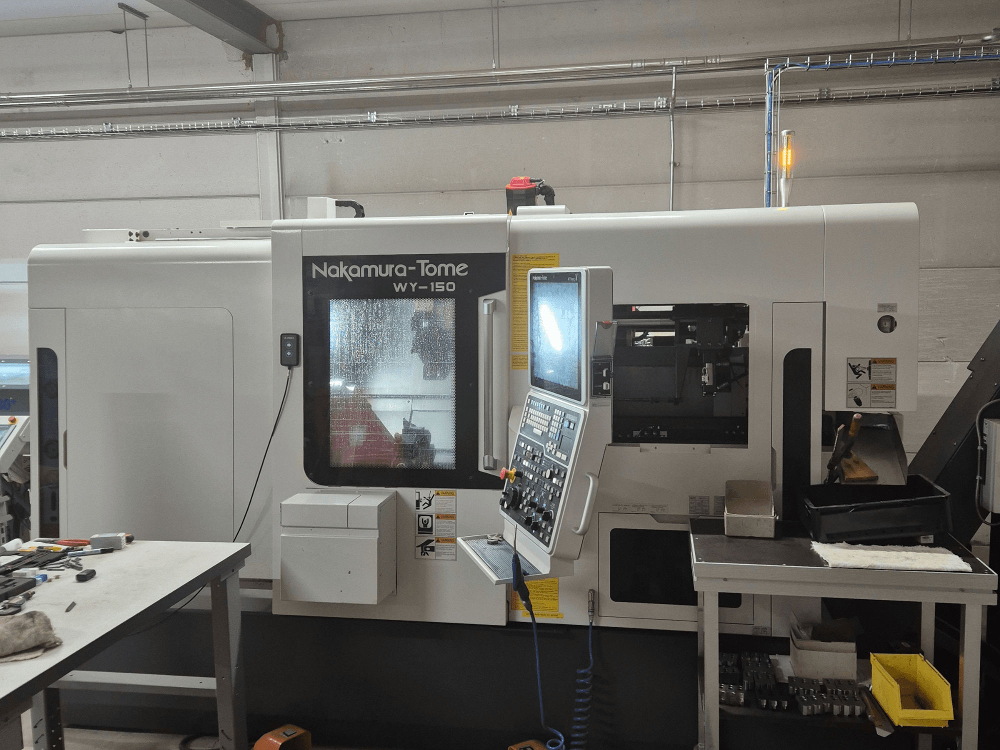 Torno CNC Nakamura-Tome WY-150 em vista lateral, com painel de controlo, janela transparente e área de trabalho visível.