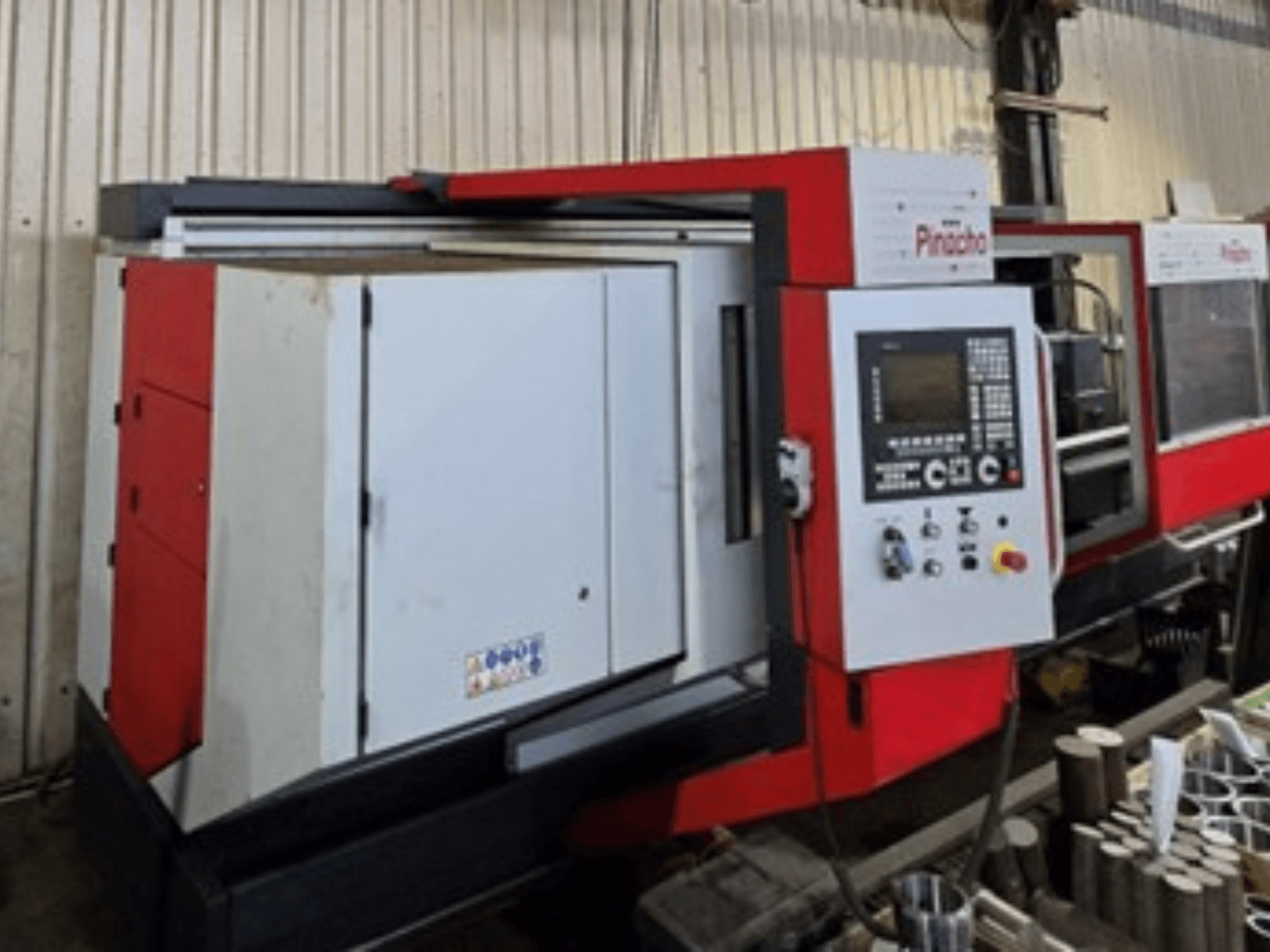 Máquina CNC Pinacho em vermelho e branco, vista lateral, com painel de controlo e materiais no chão, mostrando a configuração operacional.