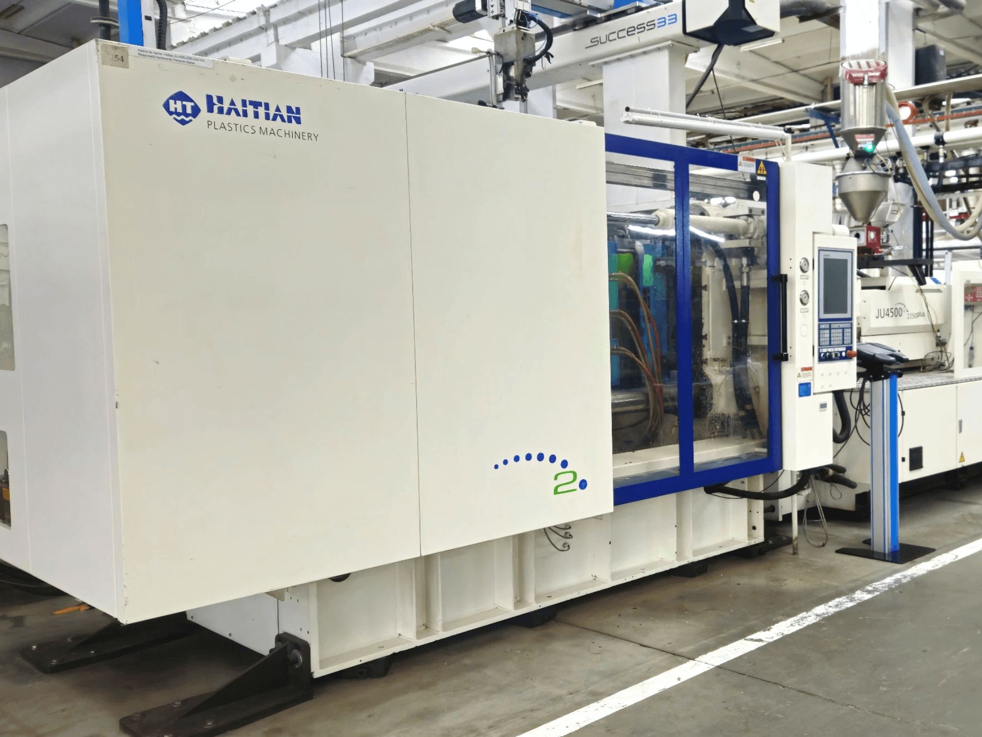 Modelo da Haitian Plastics Machinery, vista frontal, mostrando a máquina com painel de controlo e tubagem visível, num ambiente industrial.