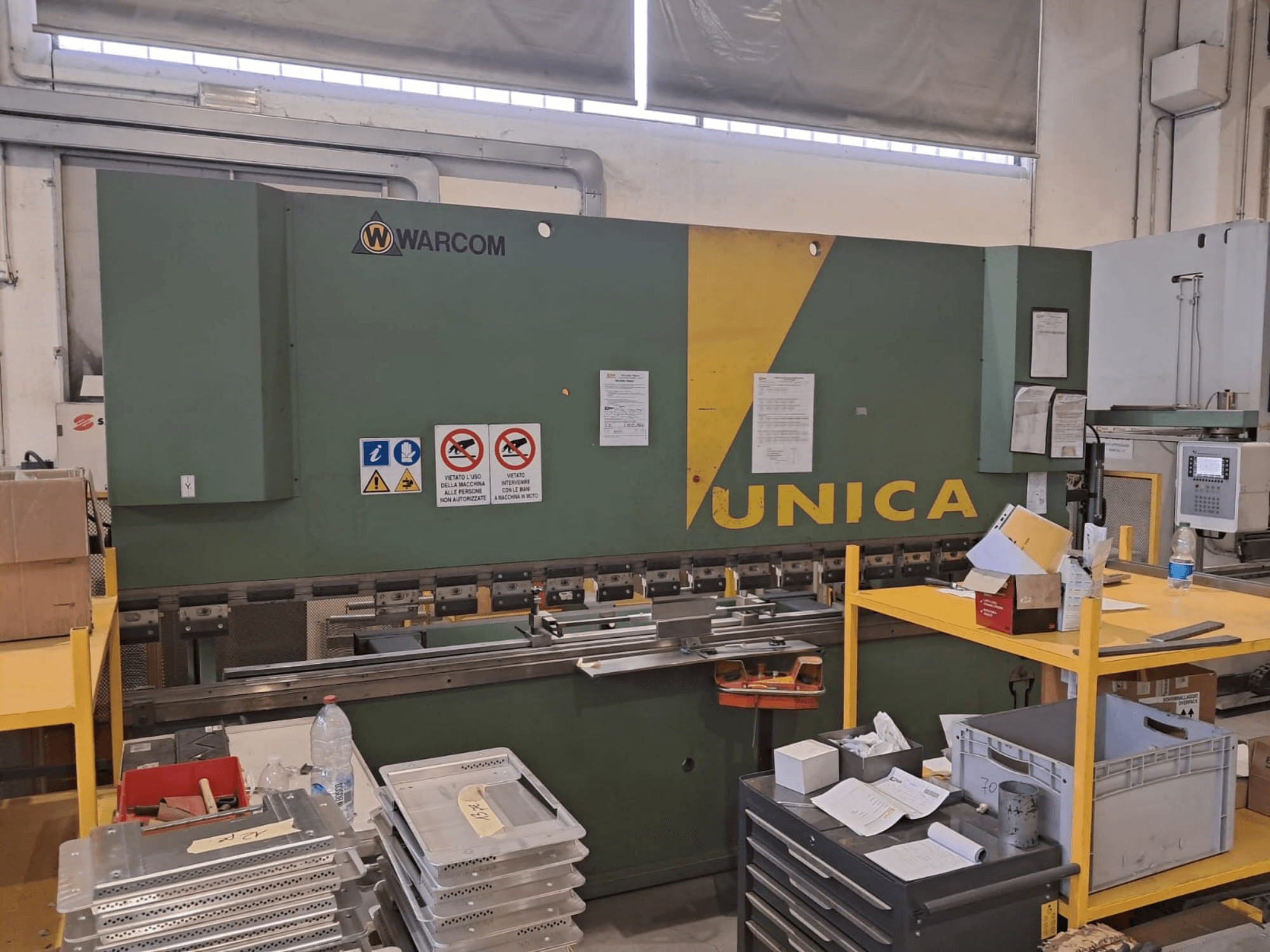 Prensa hidráulica Warcom UNICA 100-40, vista frontal, corpo verde com detalhes a amarelo, sinais de segurança visíveis, ferramentas e materiais na mesa.
