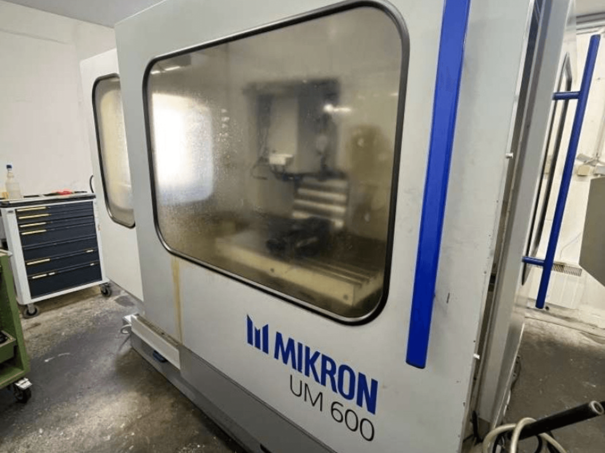 Centro de maquinagem universal MIKRON UM 600 TNC 410/426, vista lateral, com vidro embaciado e armazenamento de ferramentas visível na proximidade.