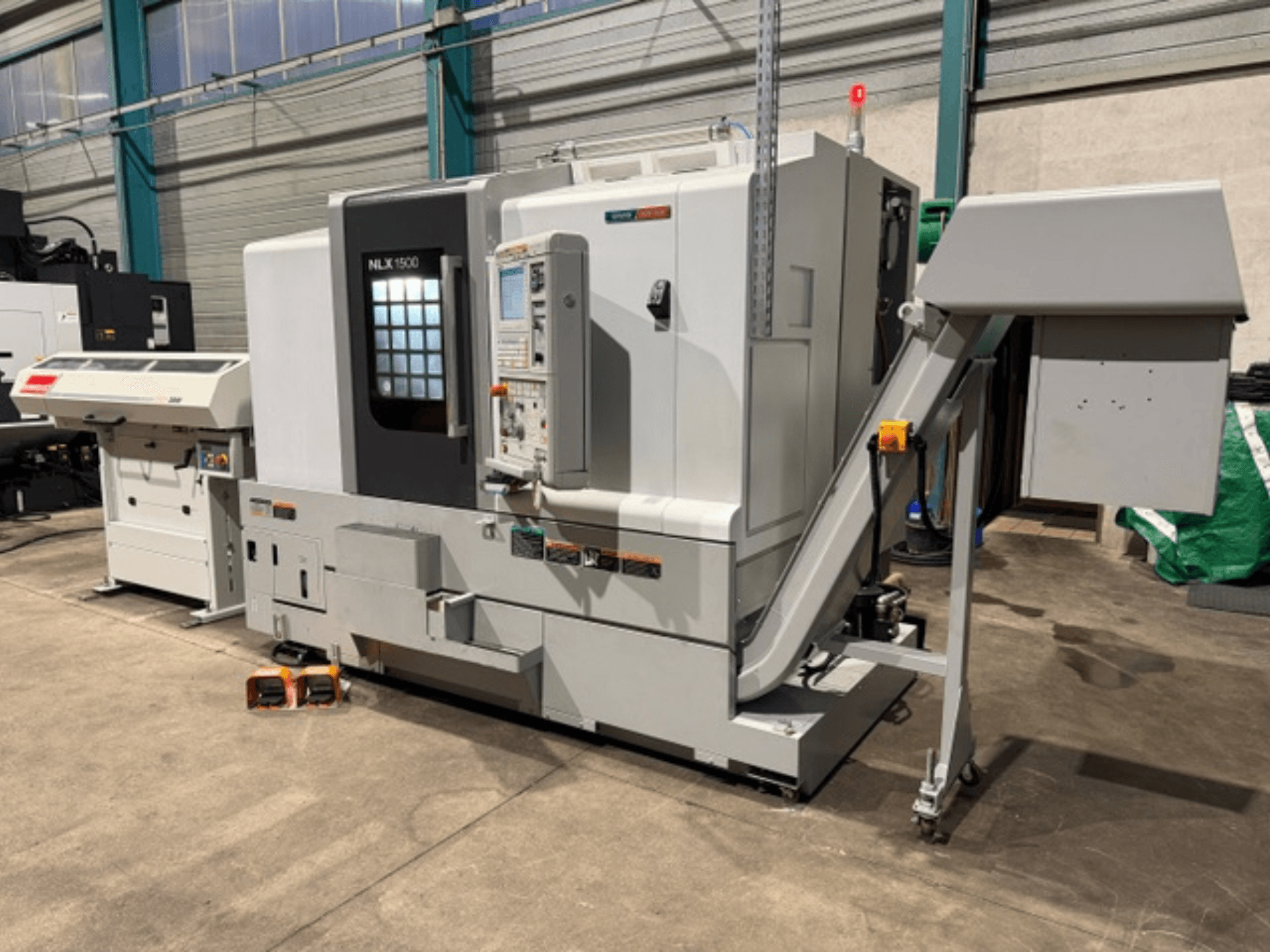 Máquina CNC NLK 1500, vista frontal, com painel de controlo e funil de acessórios ao lado, assente em chão de betão.