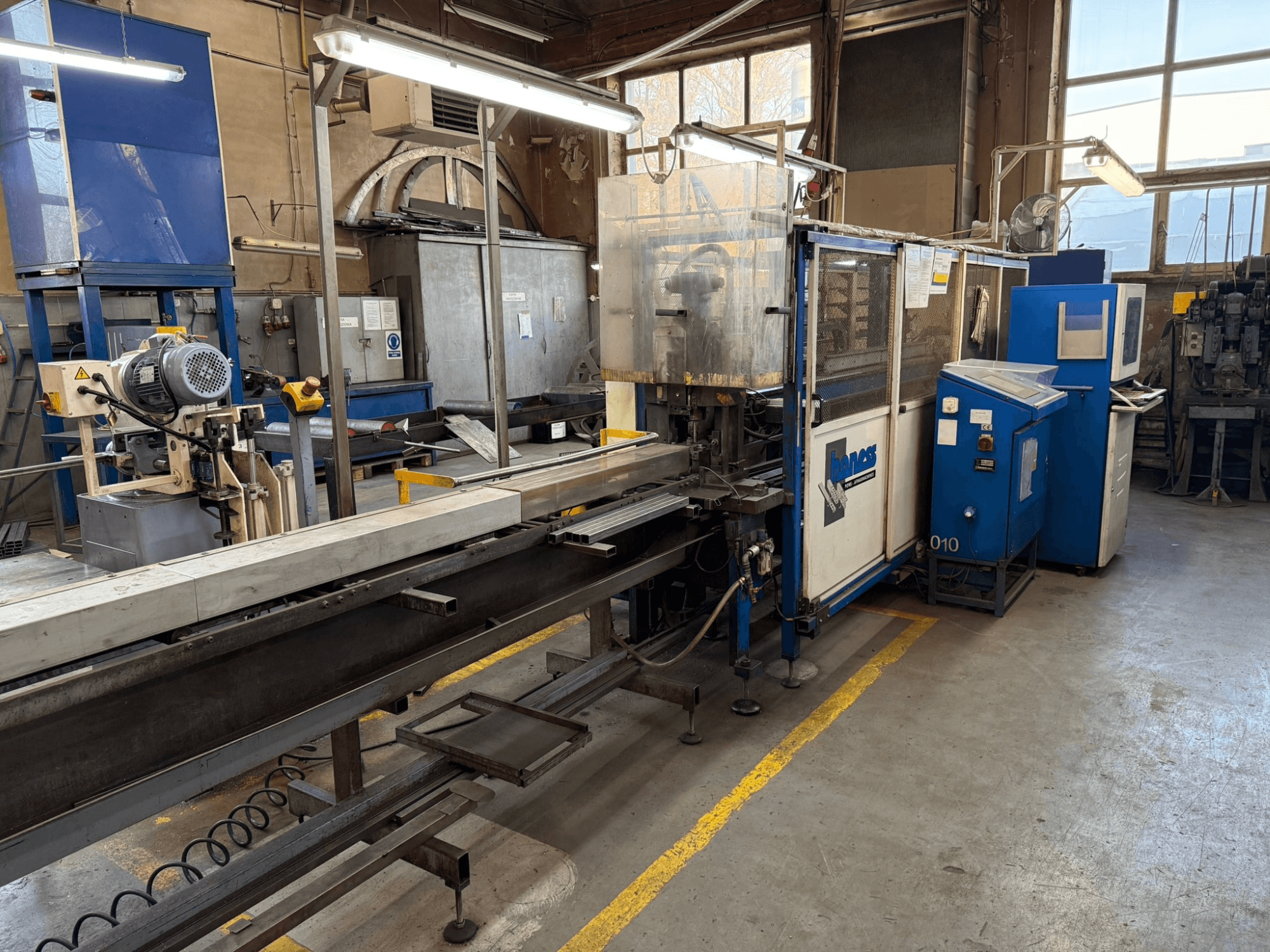 Oficina industrial com uma máquina Haweas azul, vista lateral mostrando o transportador e o equipamento num espaço grande e bem iluminado.