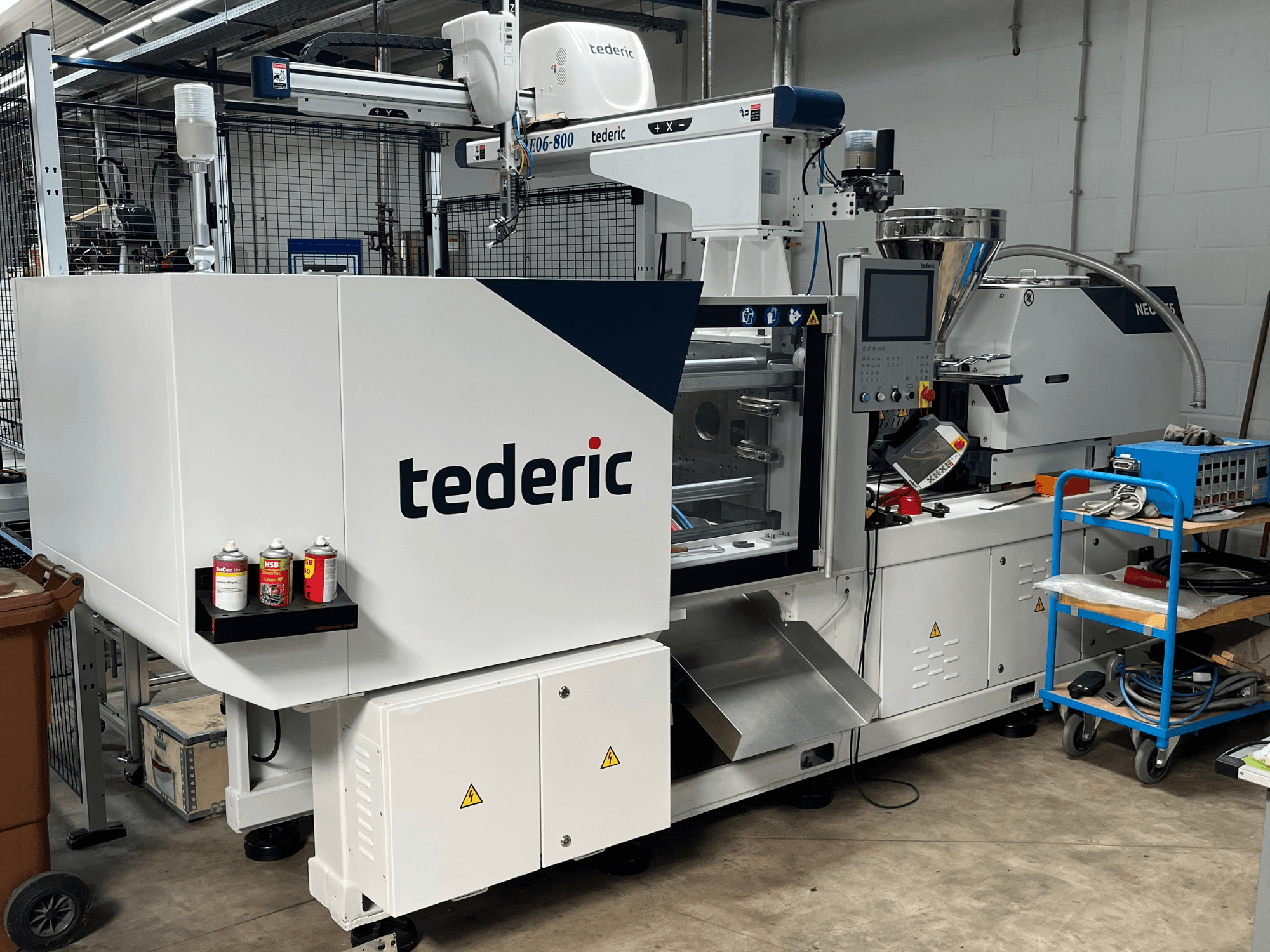 Máquina de moldagem por injeção hidráulica TEDERIC NEO.E55/e110h numa oficina, vista lateral com o painel de controlo e as ferramentas adjacentes.
