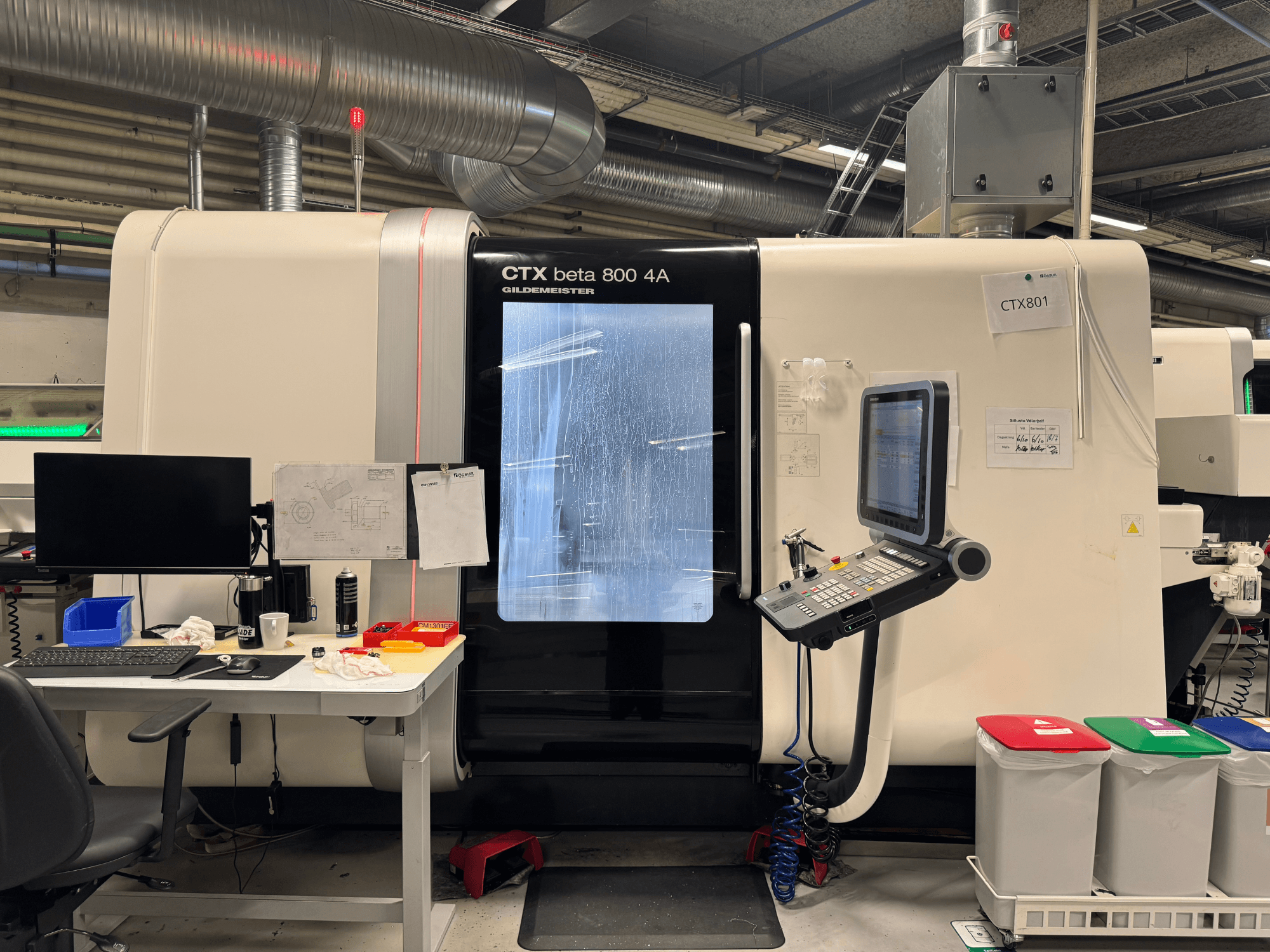 Máquina CTX beta 800 4A com painel de controlo e monitor, vista lateral, numa oficina com configuração de escritório e caixotes do lixo coloridos.
