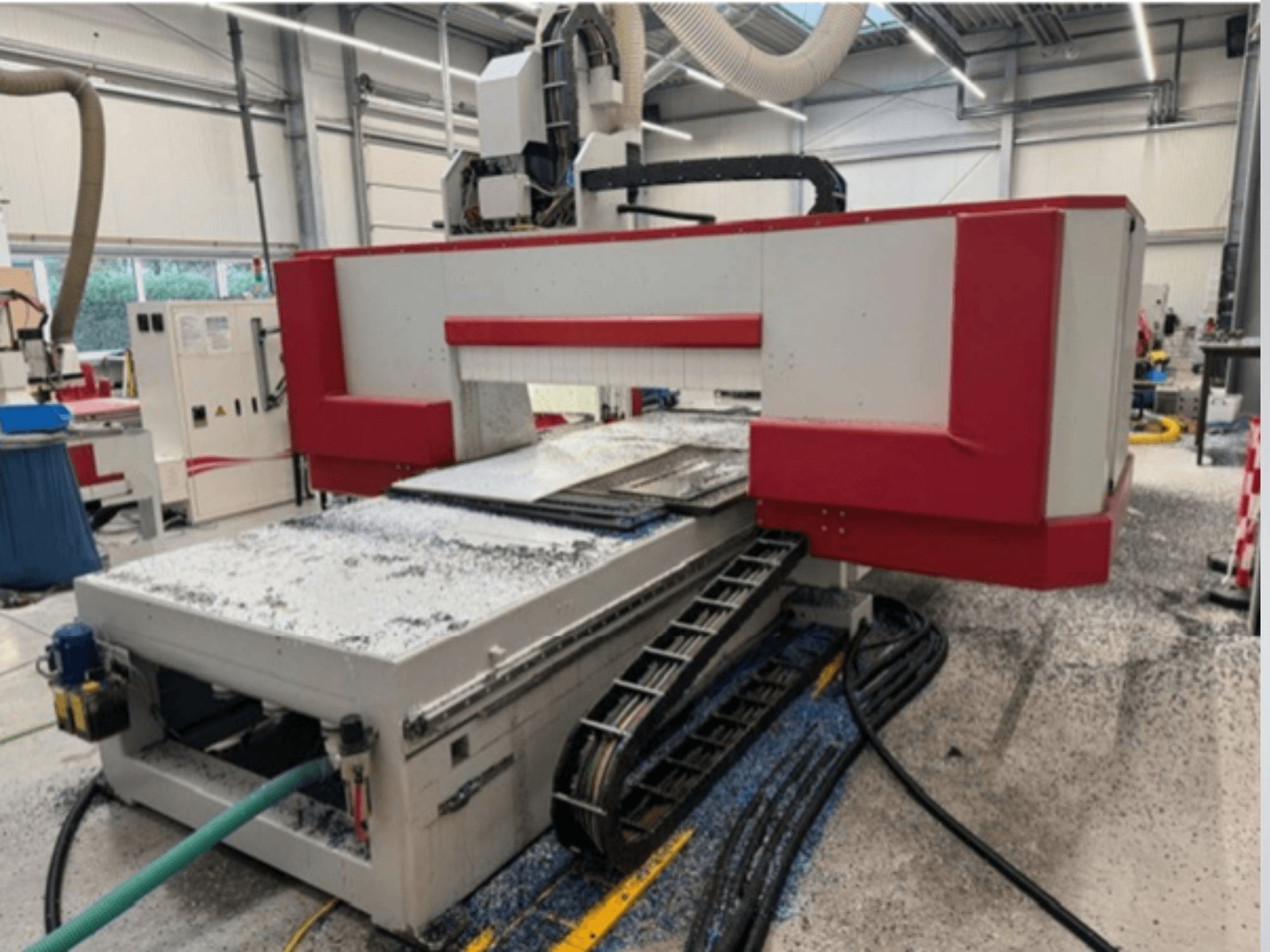 Máquina CNC vermelha e cinzenta numa oficina, vista lateral mostrando a mesa de materiais, a mangueira de refrigeração e a serradura no chão.