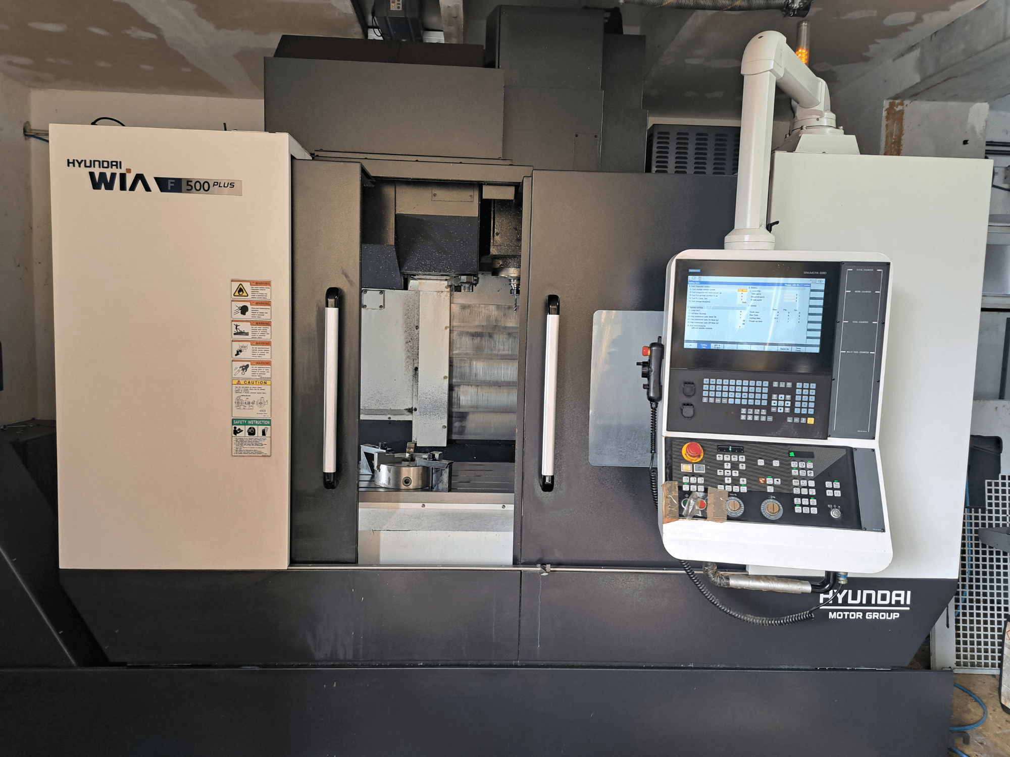 Máquina CNC Hyundai WIA F500 Plus em vista frontal, com um painel de controlo digital e etiquetas de segurança.