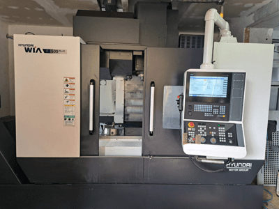 Máquina CNC Hyundai WIA F500 Plus em vista frontal, com um painel de controlo digital e etiquetas de segurança.