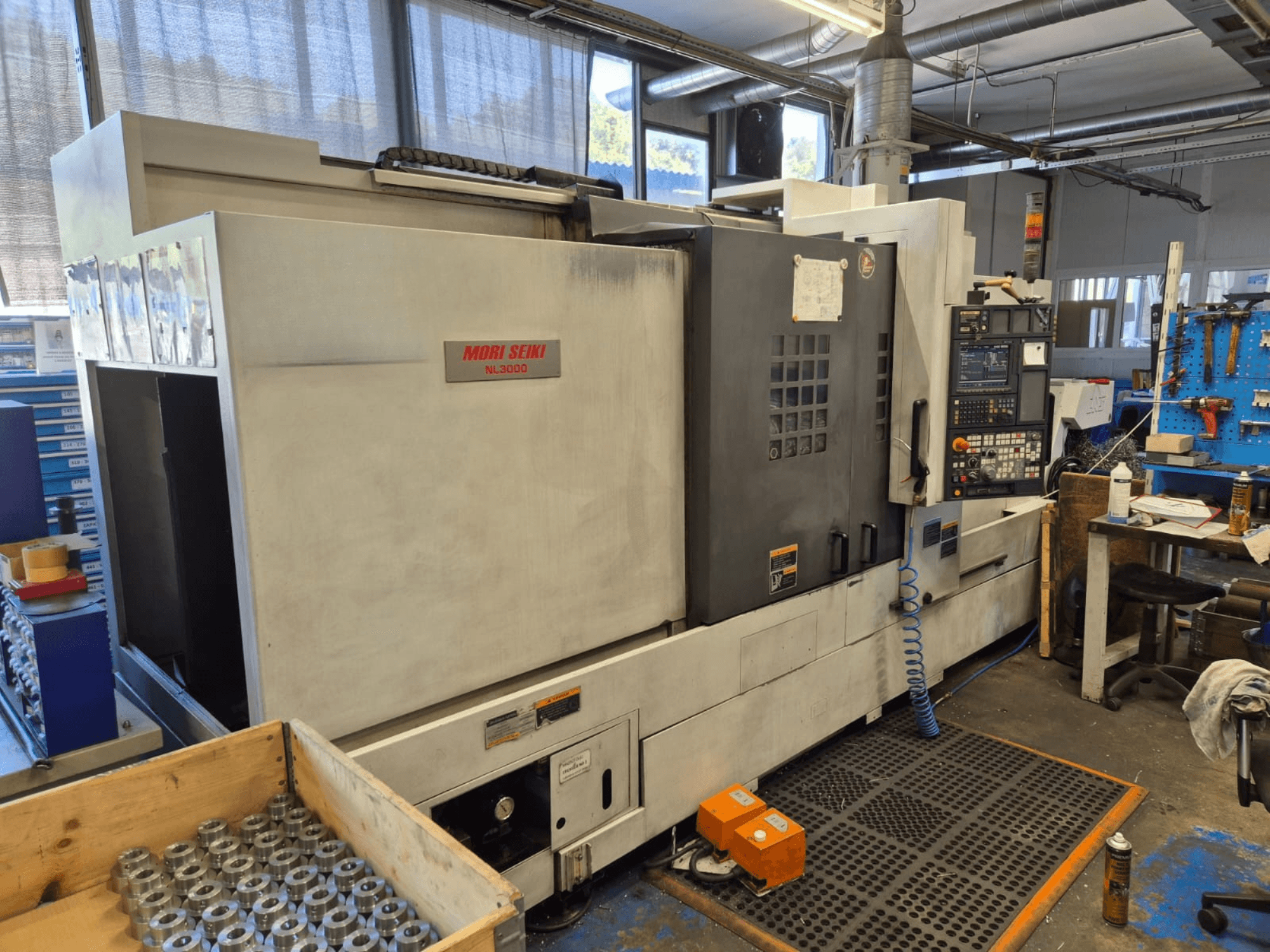 Torno CNC Mori Seiki NL3000 numa oficina, vista lateral mostrando o painel de controlo, ferramentas e peças metálicas numa caixa de madeira.