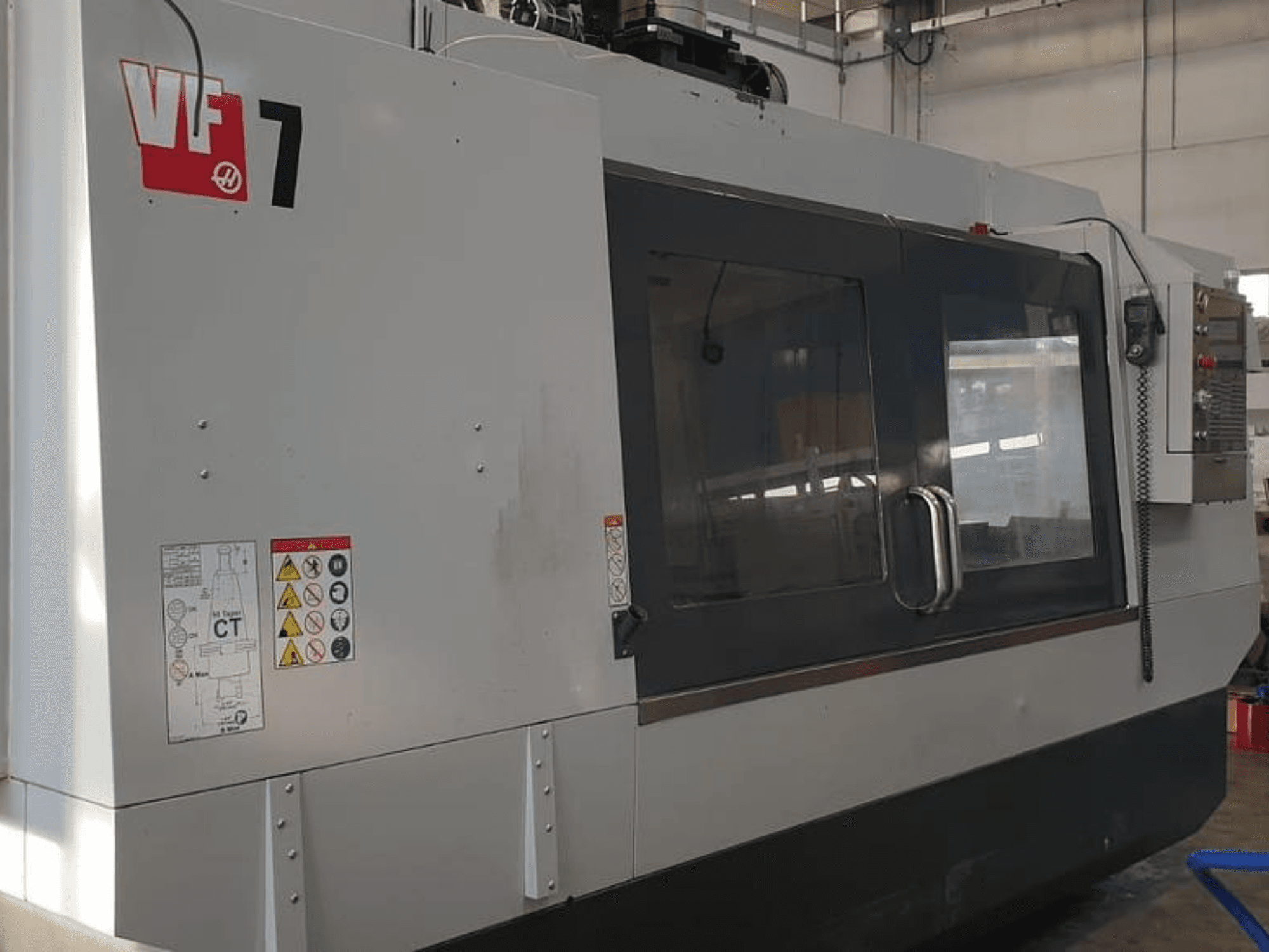 Máquina CNC Haas VF-7 em vista lateral, apresentando um acabamento em cinzento metálico, painel de controlo e etiquetas de segurança.