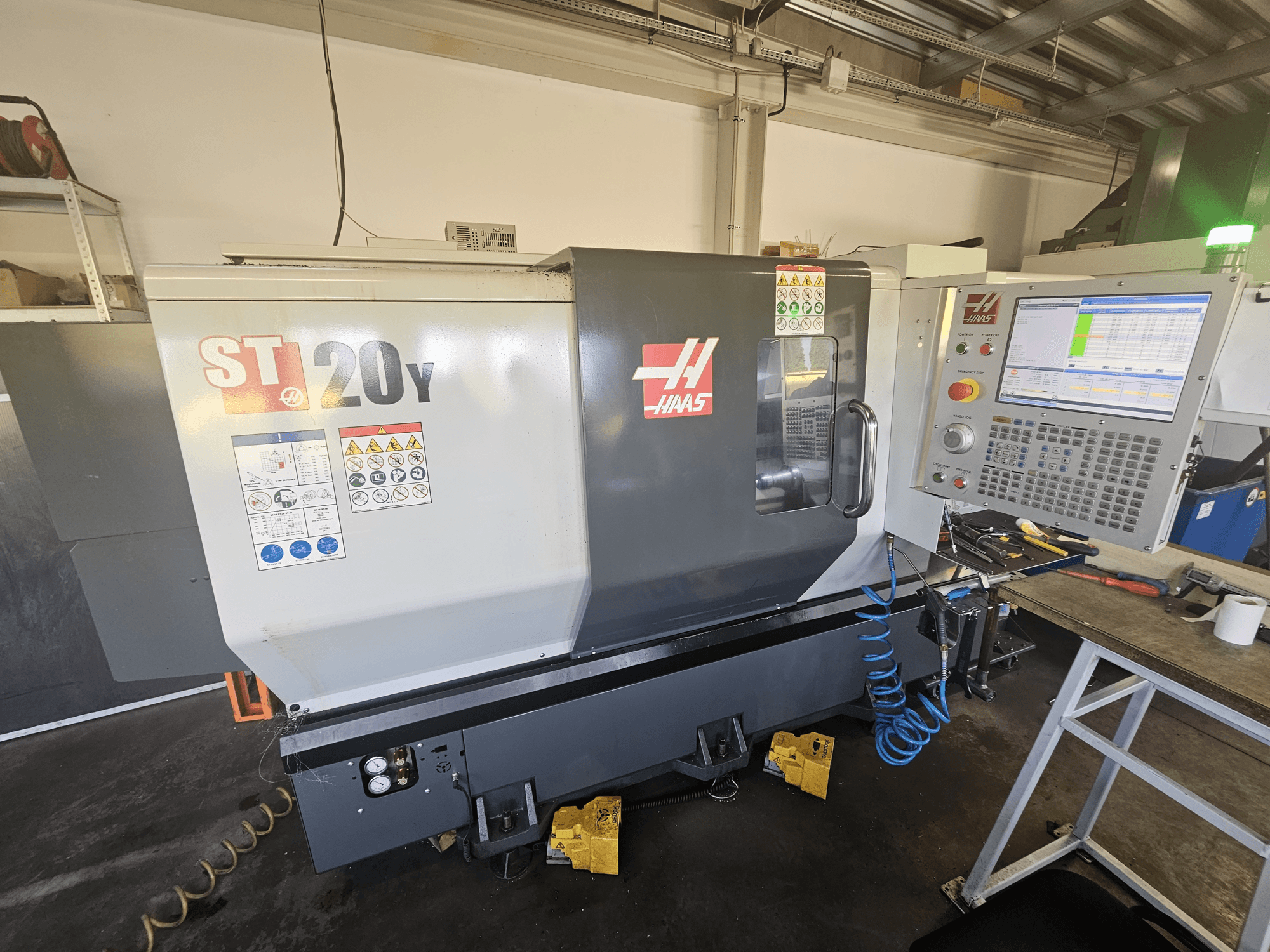 Torno CNC Haas ST-20Y em vista frontal com painel de controlo; inclui etiquetas de segurança e detalhe da mangueira de ar na lateral.
