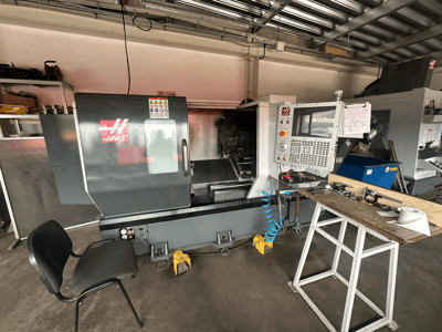 Torno CNC Haas (modelo ST-10) numa oficina, vista lateral mostrando os controlos, ferramentas numa bancada de trabalho e uma cadeira próxima.