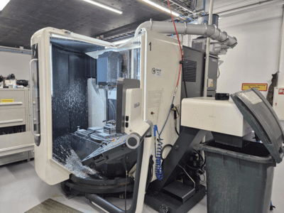 Fresadora DMG aberta com pulverização de líquido de refrigeração, com aparas de metal e comandos de ferramentas, vista de frente.