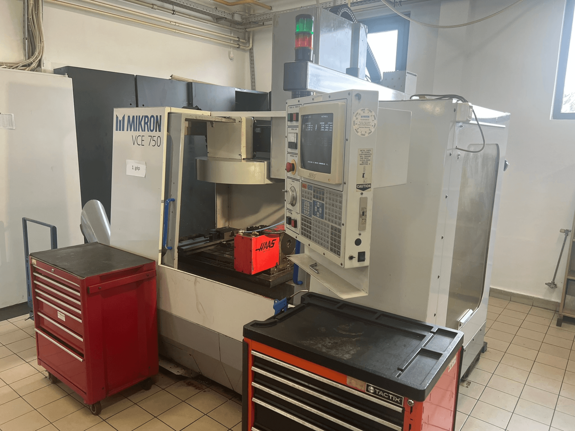 Texto alternativo: "Centro de maquinação vertical MIKRON VCE 750, vista frontal com painel de controlo e caixas de ferramentas vermelhas, numa oficina espaçosa."