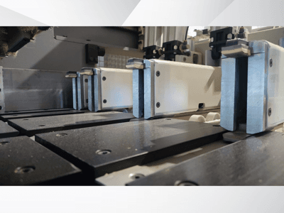 Vista em grande plano do mecanismo de fixação de uma máquina de precisão com maxilas metálicas, com superfícies de trabalho pretas e calhas de guia visíveis.