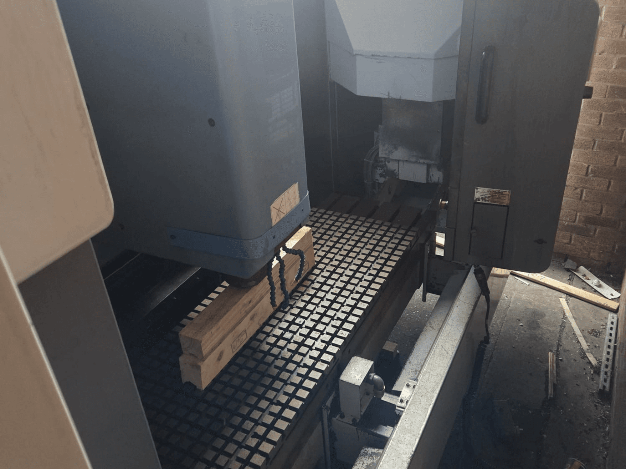 Vista frontal de uma máquina CNC com pranchas de madeira numa mesa de grelha, mostrando ferramentas e controlos na lateral.