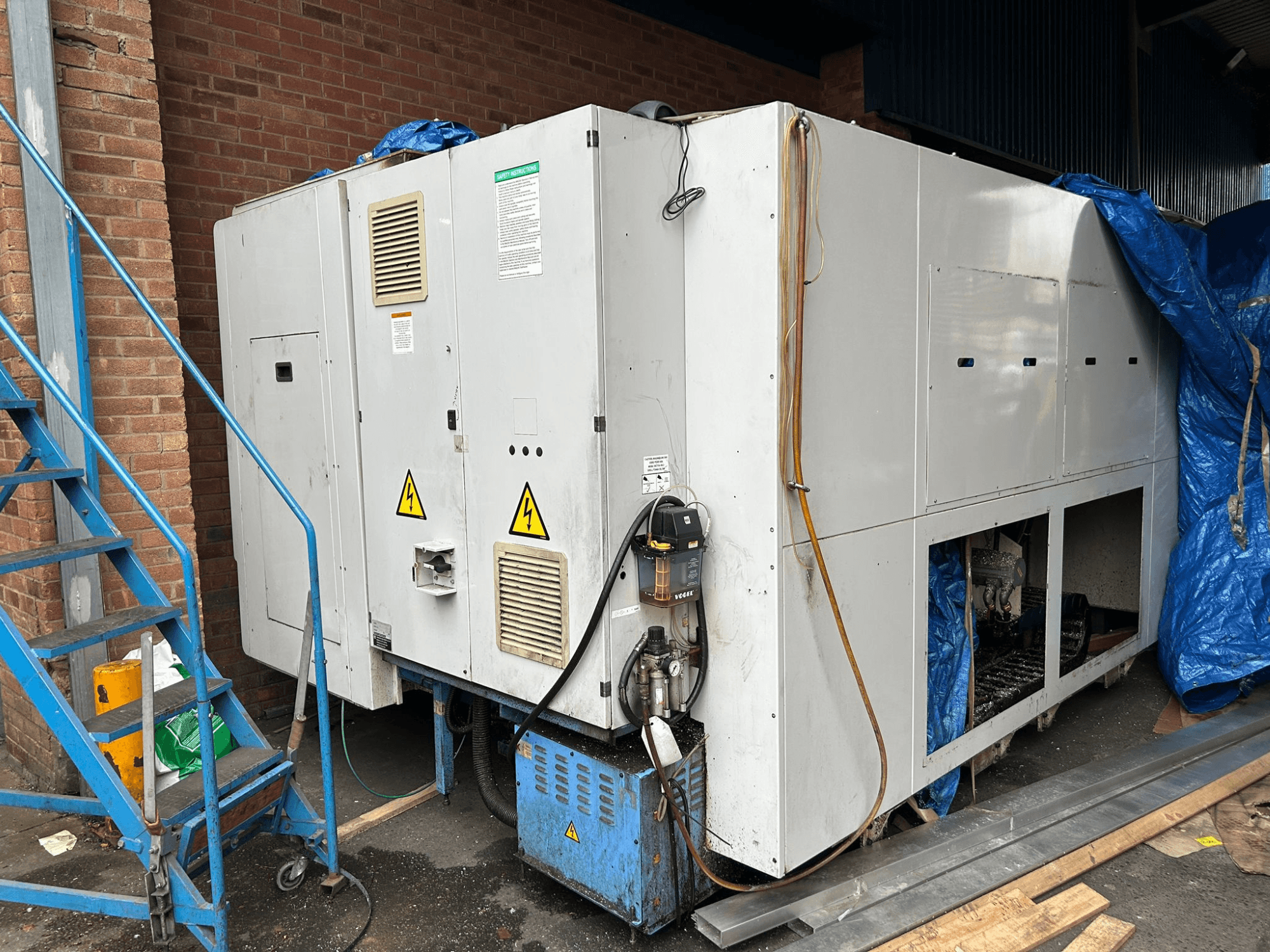 Vista lateral de um grande refrigerador industrial, de marca e modelo pouco claros, mostrando ligações eléctricas, sinais de aviso e cobertura de lona azul.