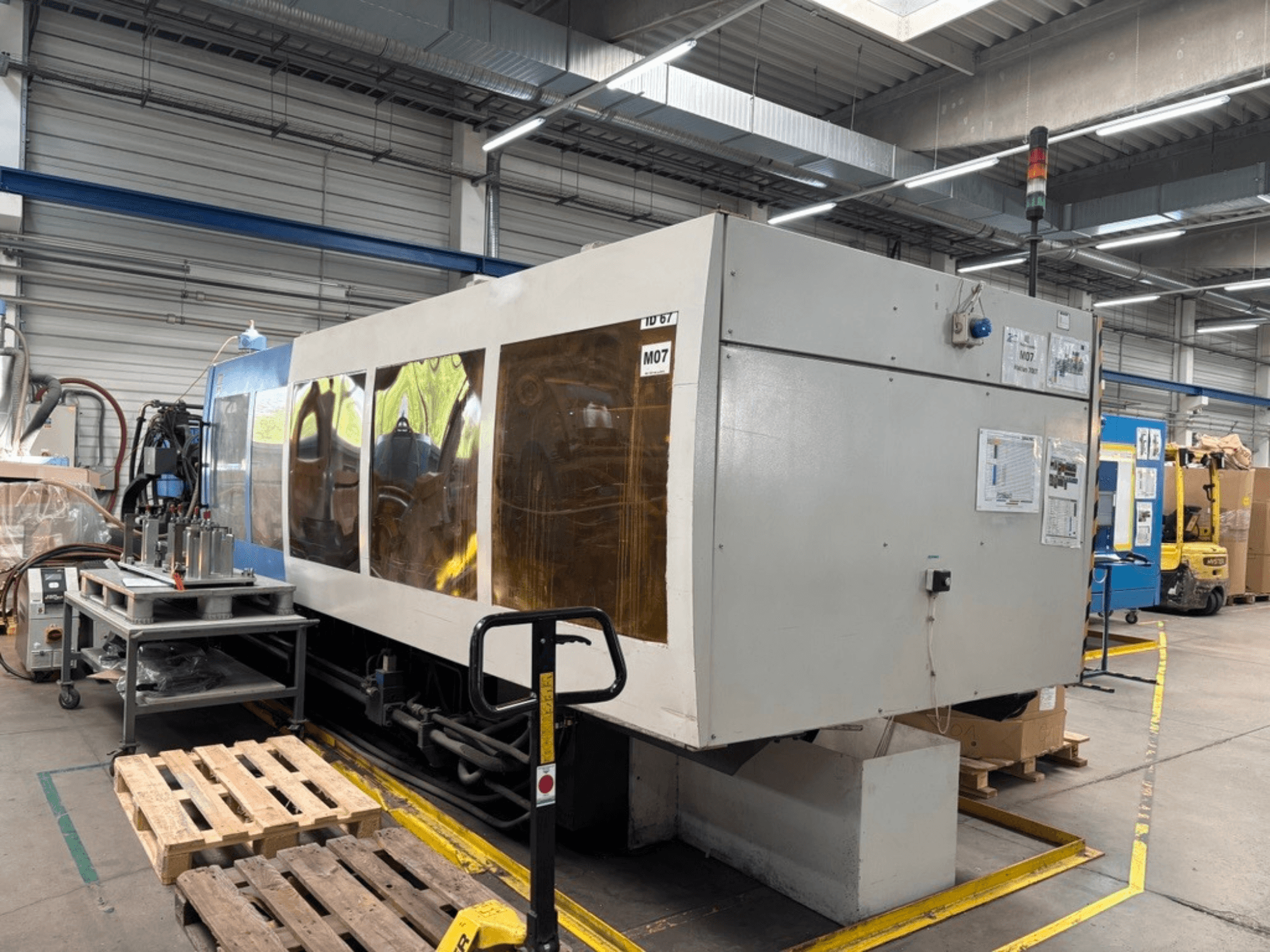 Máquina de moldagem por injeção hidráulica HAITIAN 7000/5000 numa fábrica, vista lateral com painéis de controlo e luzes de segurança visíveis.