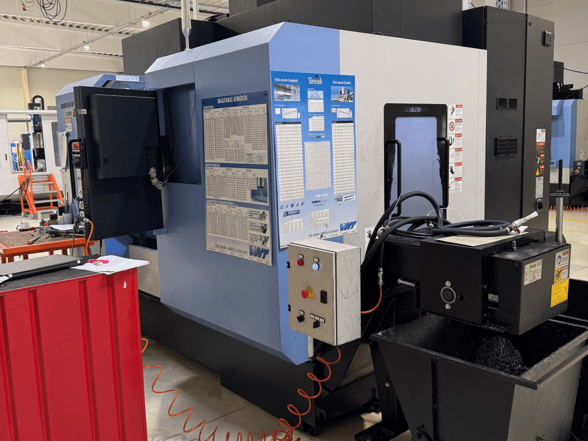 Centro de fresagem e torneamento DOOSAN DNM350/5AX, vista lateral que mostra o painel de controlo e o depósito de líquido de arrefecimento, com detalhes operacionais visíveis.