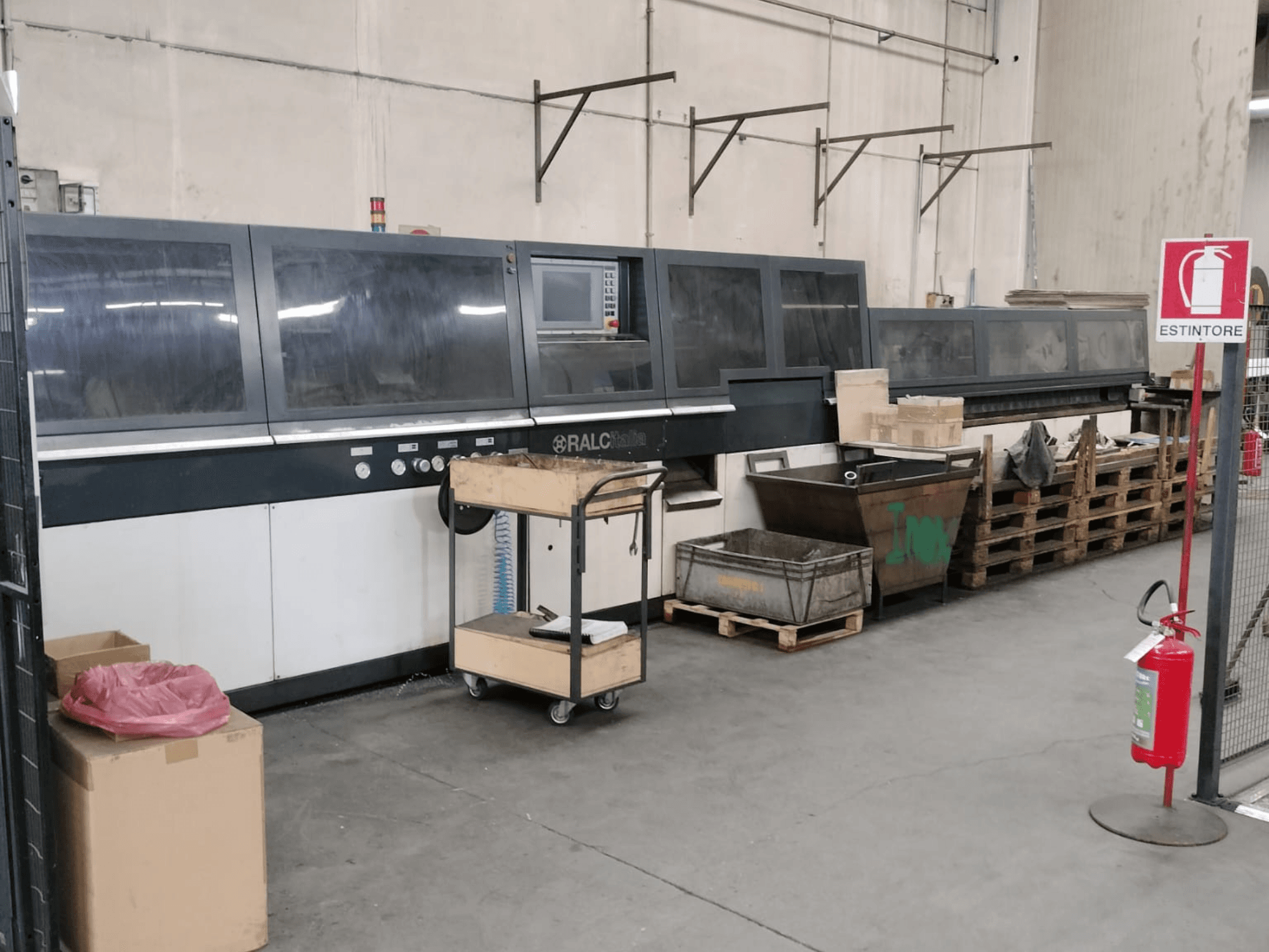 Máquina industrial RALC em ambiente fabril, vista frontal, com painel de controlo visível; paletes de madeira e extintor de incêndio nas proximidades.