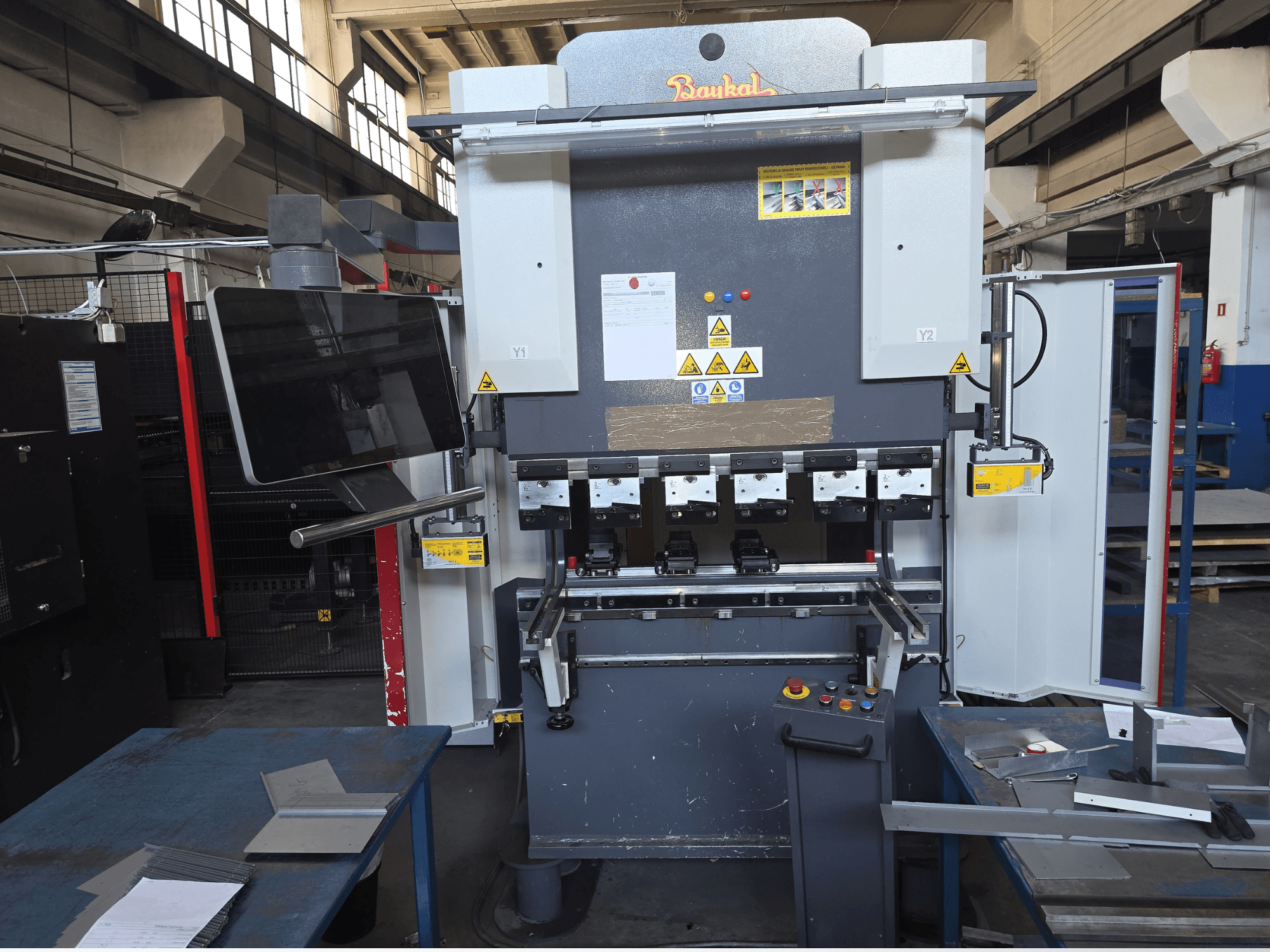 Prensa-dobra Baykal APHS 125040, vista frontal, mostrando o painel de controlo e os dispositivos de segurança, com materiais sobre uma mesa de trabalho azul.