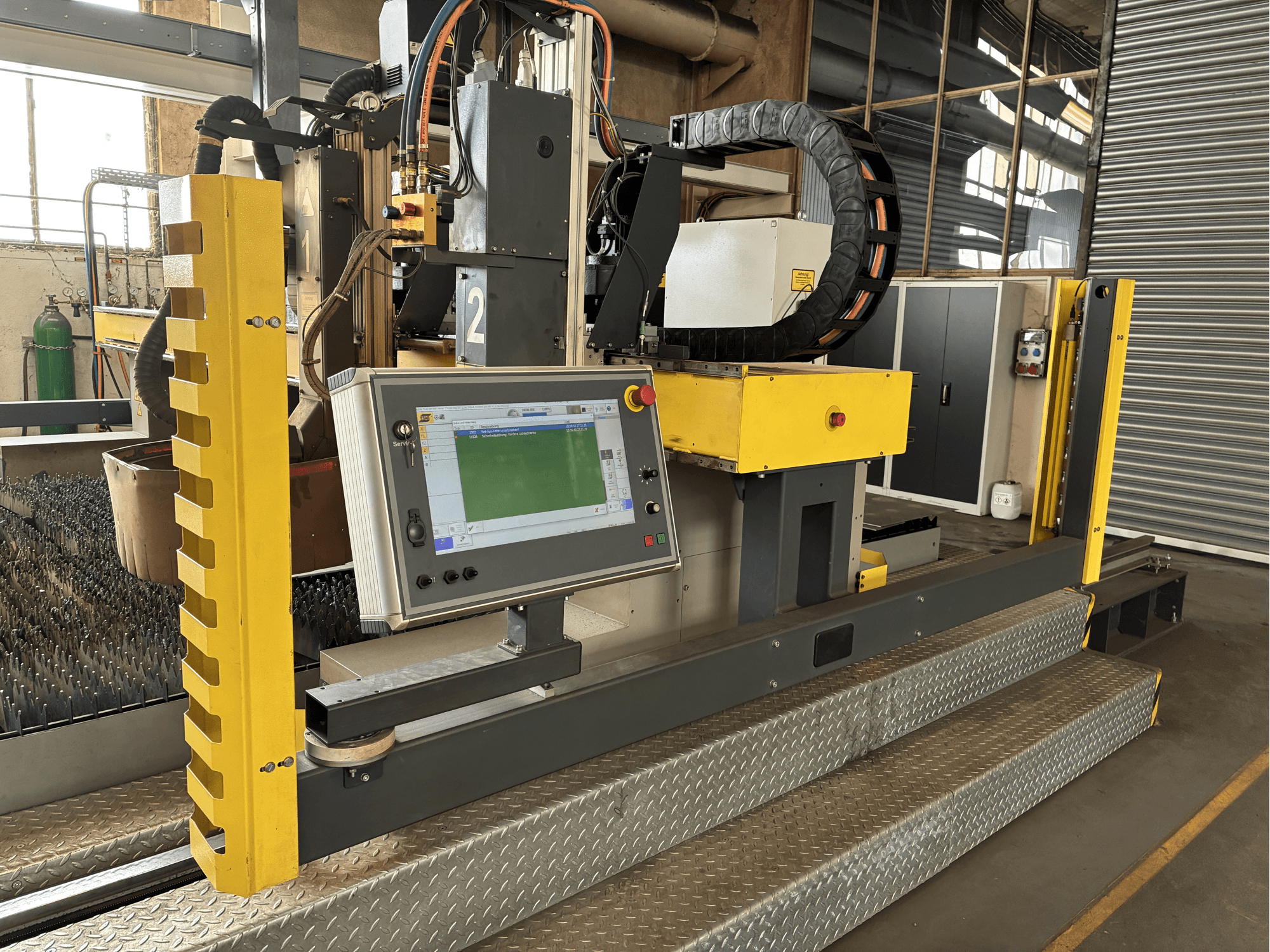 Máquina de corte industrial com painel de controlo com ecrã tátil, proteção de segurança amarela, vista frontal, base de grelha metálica visível.