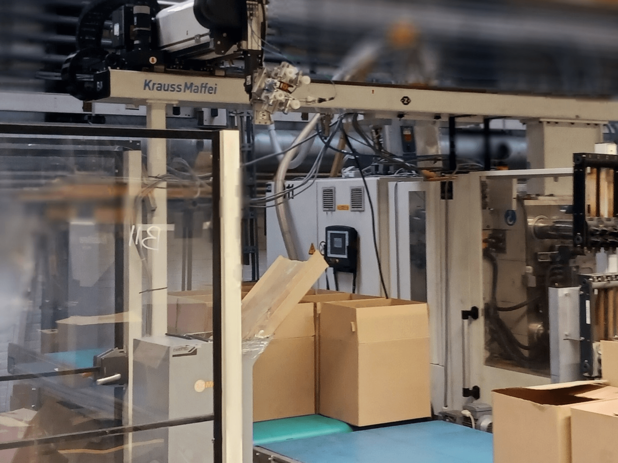 Sistema de automação Krauss Maffei numa fábrica, vista frontal mostrando caixas num transportador e componentes de máquinas em funcionamento.