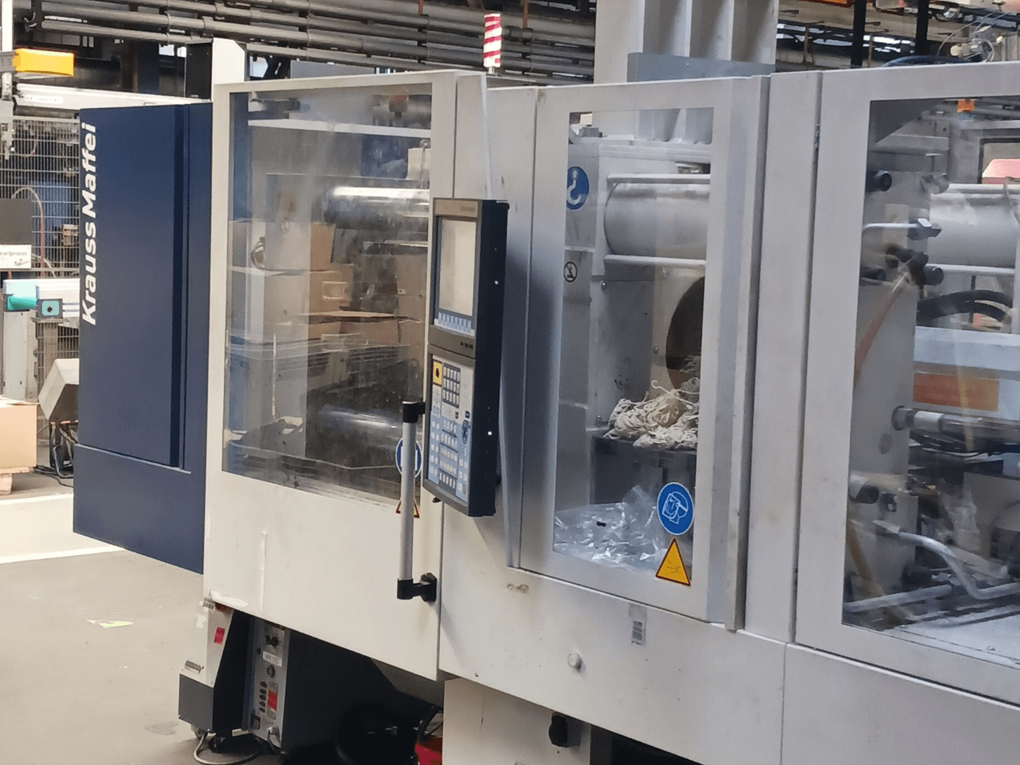 Máquina de moldagem por injeção KraussMaffei, vista lateral, mostrando o painel de controlo, a maquinaria e os materiais de embalagem em segundo plano.