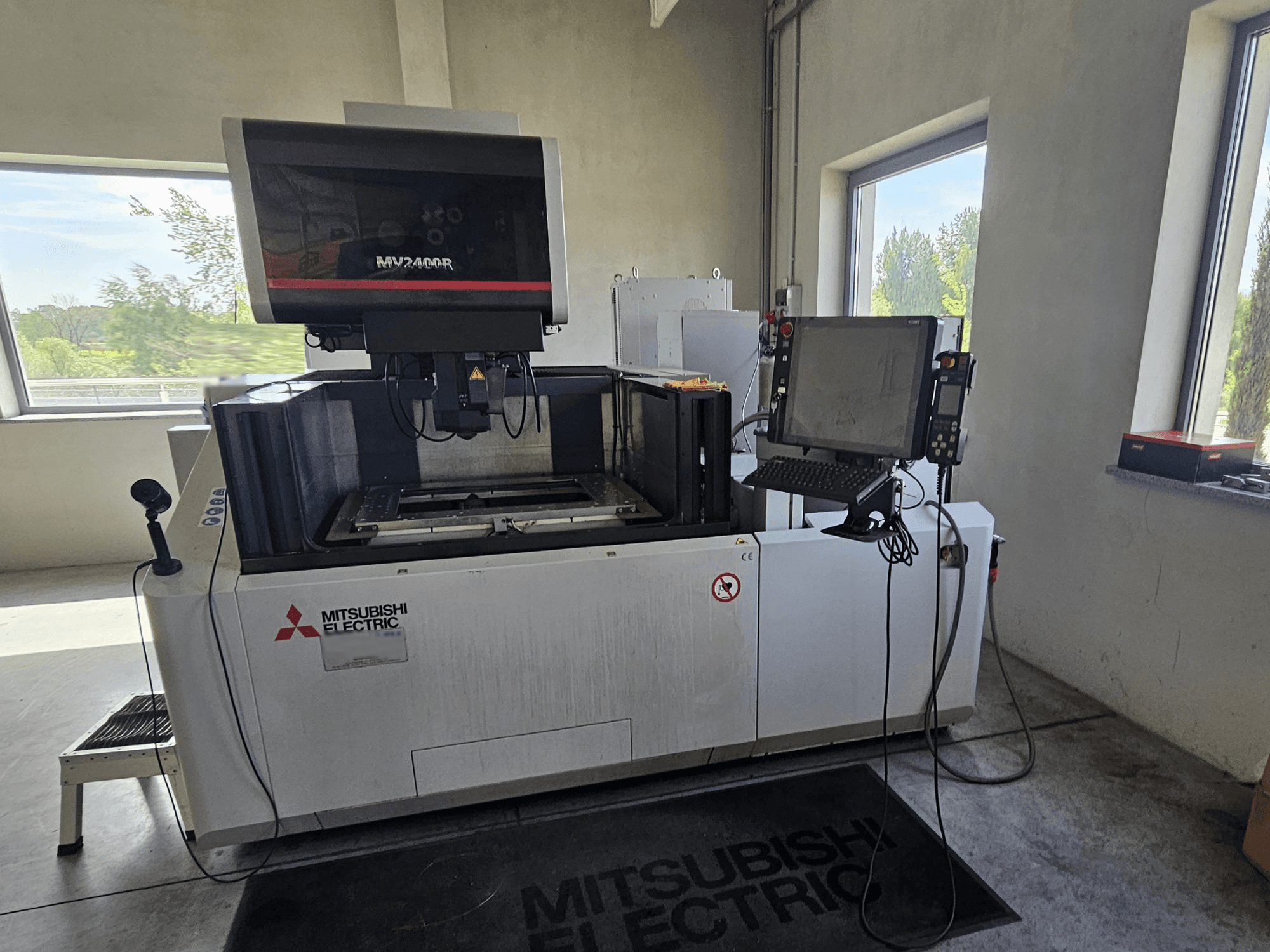 Máquina EDM de fio MV2400R da Mitsubishi numa oficina, com um painel de controlo com ecrã tátil e uma área de trabalho.