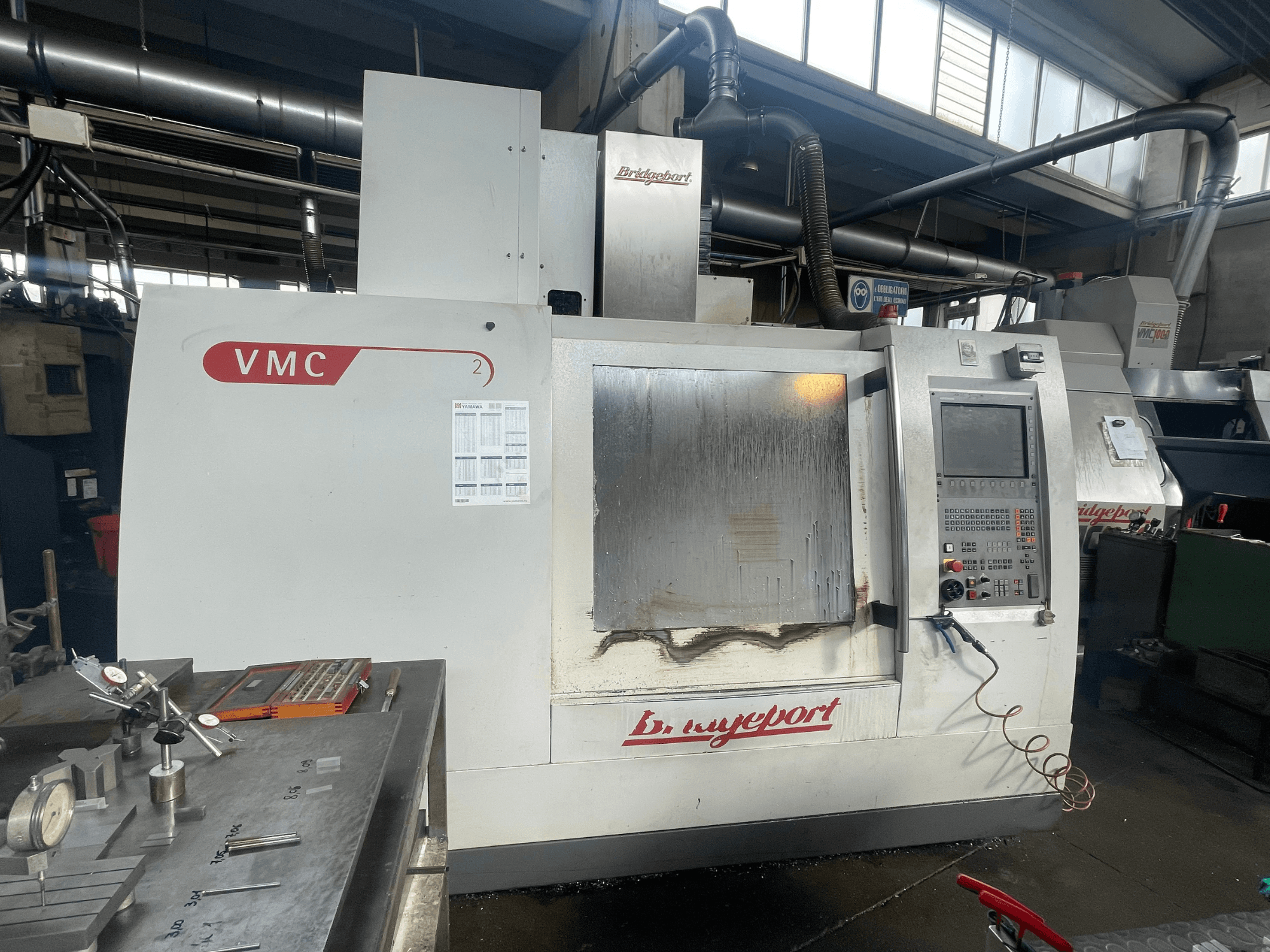 Centro de maquinagem vertical Bridgeport VCM 800-30 numa oficina, com um painel de controlo e uma mesa de ferramentas. Vista frontal com detalhes visíveis.