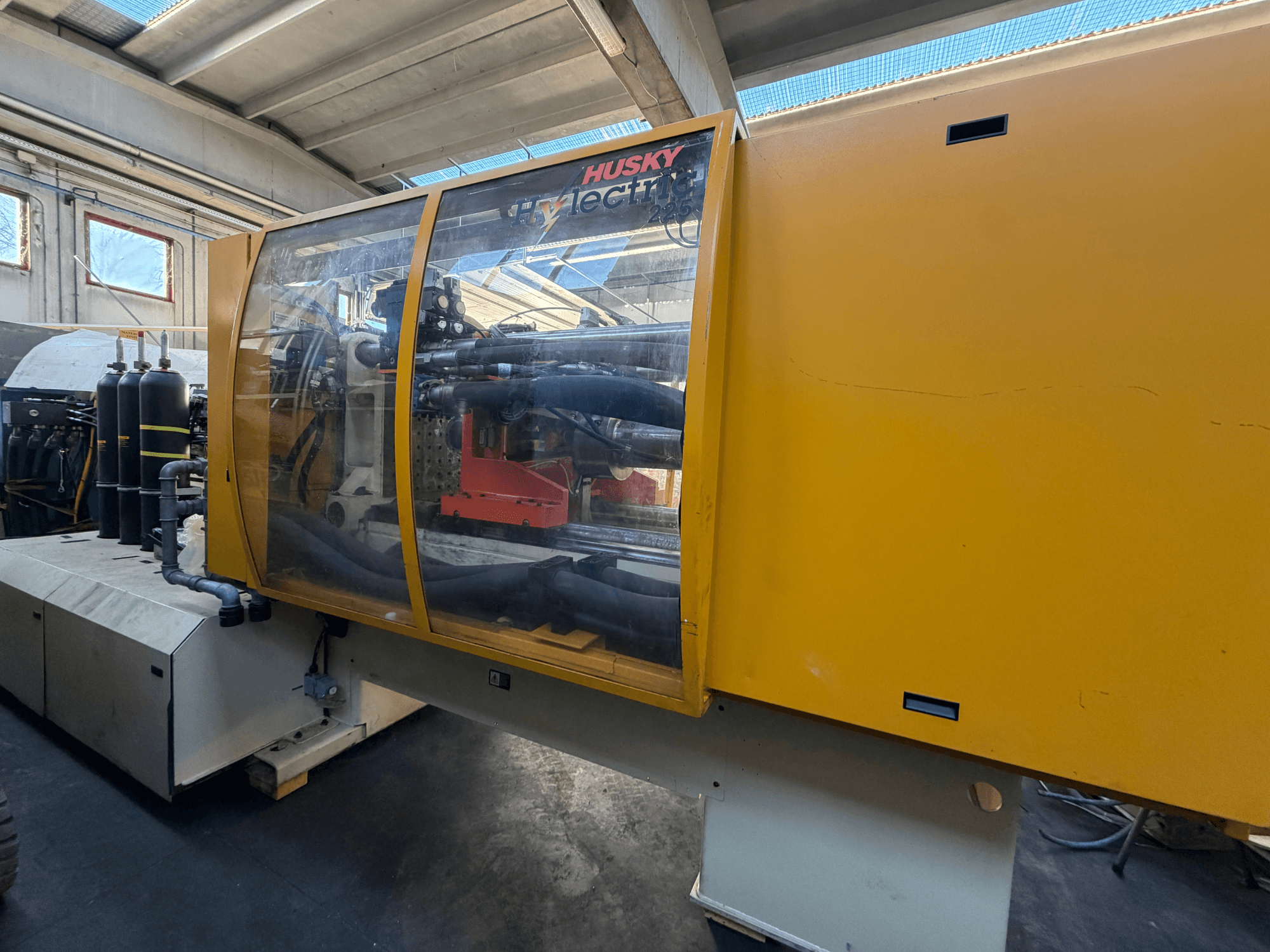 Máquina de moldagem por injeção eléctrica Husky HL225 RS65, vista frontal com caixa amarela, painel de controlo visível e componentes hidráulicos.